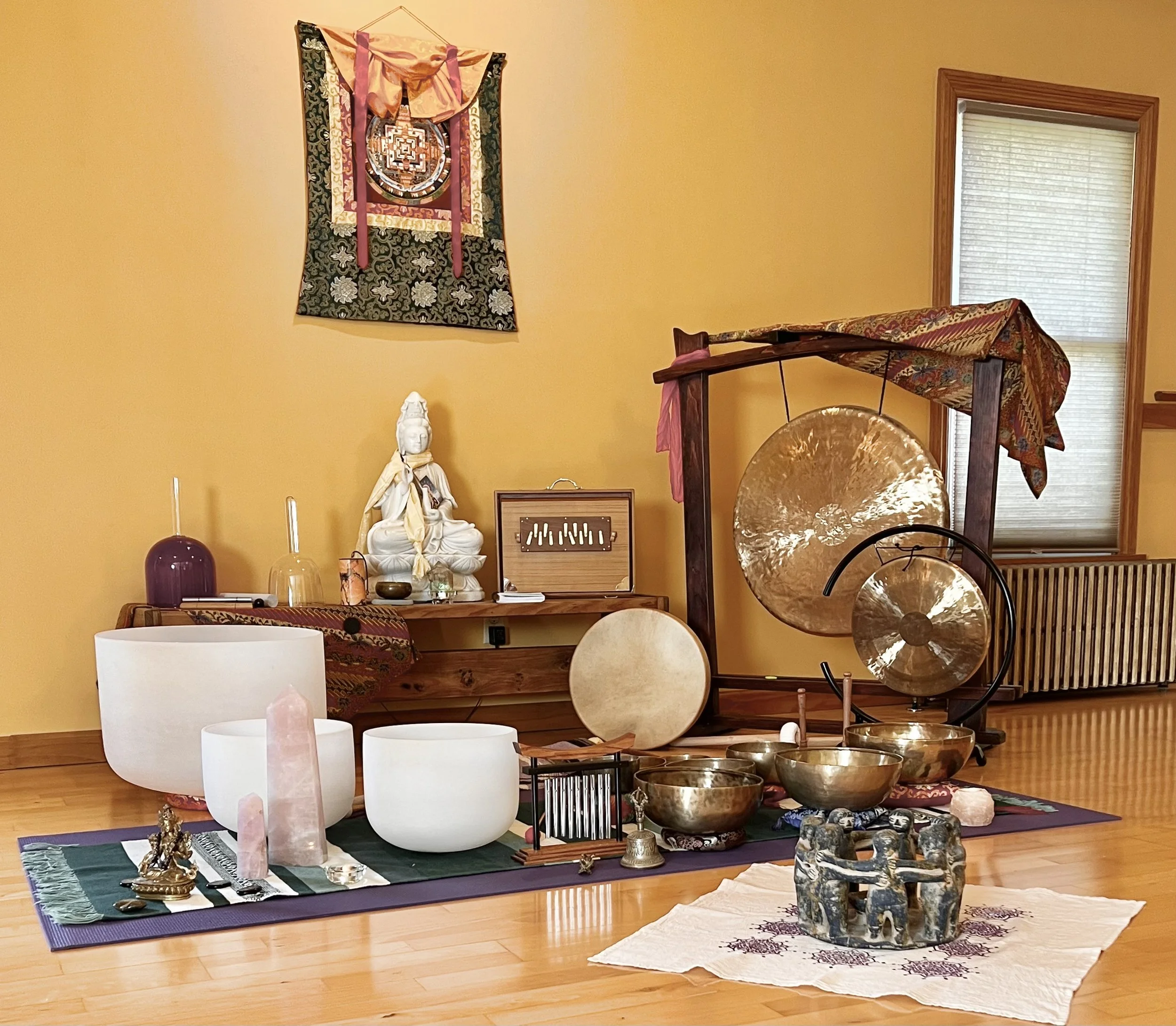 Arrangement of singing bowls, gongs, and other meditation instruments on a purple mat in a golden colored yoga studio with wooden floors, a window, and a colorful tapestry on the wall.