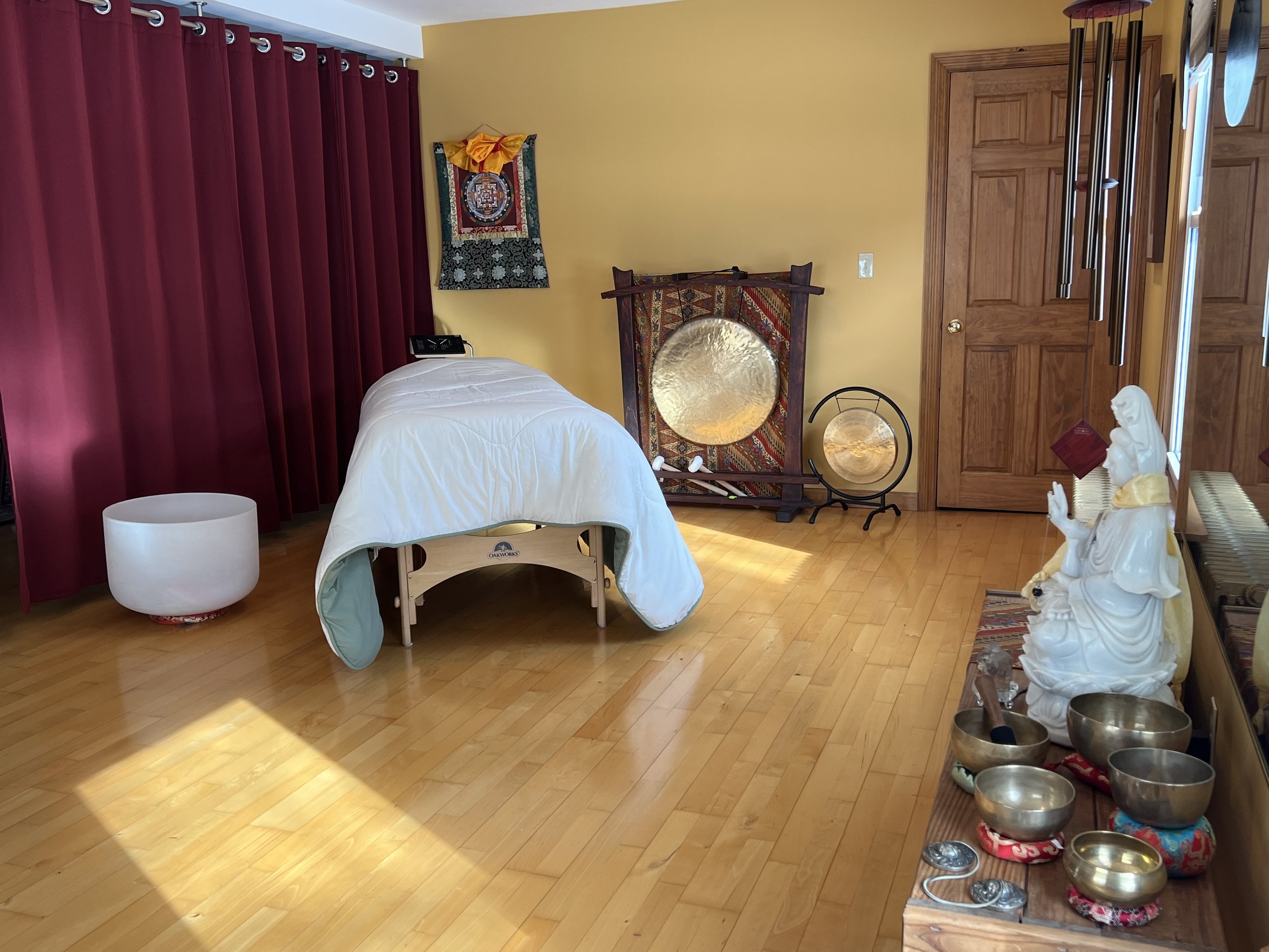 A massage table set up for a Reiki session. The room has wooden flooring, golden colored walls, and contains various decorative objects including sound healing instrumets and a marble statue of Guan Yin.