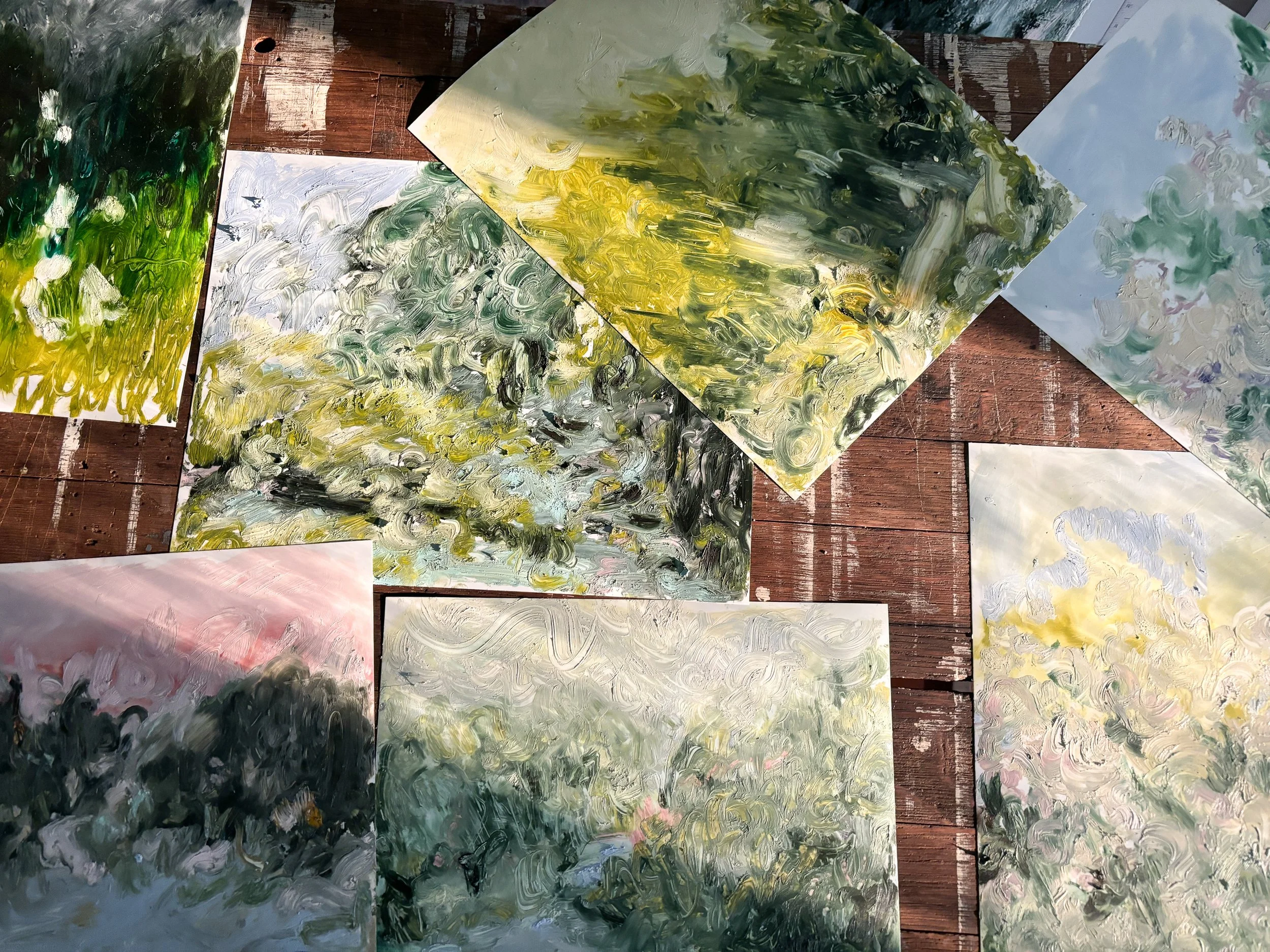 Several abstract landscape paintings on paper scattered on a wooden surface, featuring impressionistic brushstrokes in green, yellow, white, black, and light blue colors.