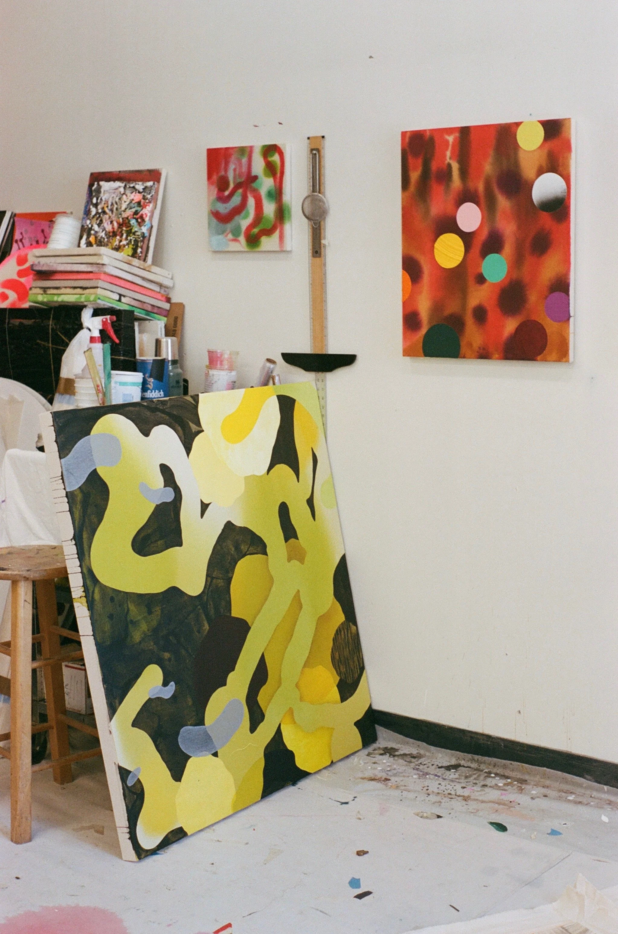 An art studio with a large abstract painting leaning against the floor, featuring yellow, black, and light blue shapes, with two smaller colorful abstract paintings on the wall, and various art supplies and stacked canvases on a table.