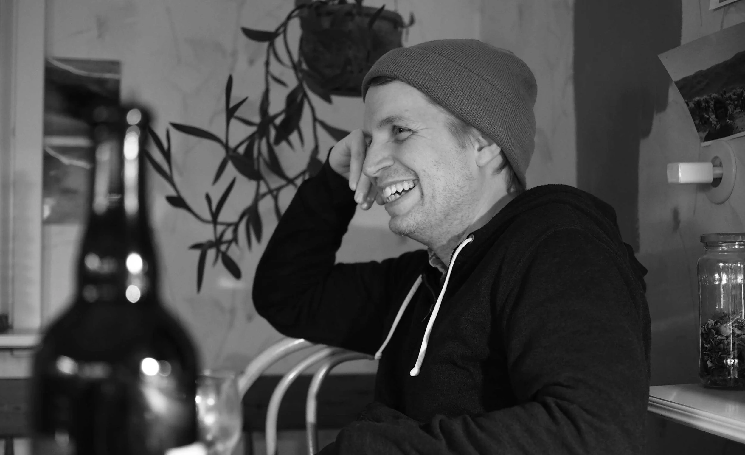 A man smiling and laughing, wearing a beanie and a hoodie, sitting at a table in a cozy indoor setting with potted plant and jars in the background.