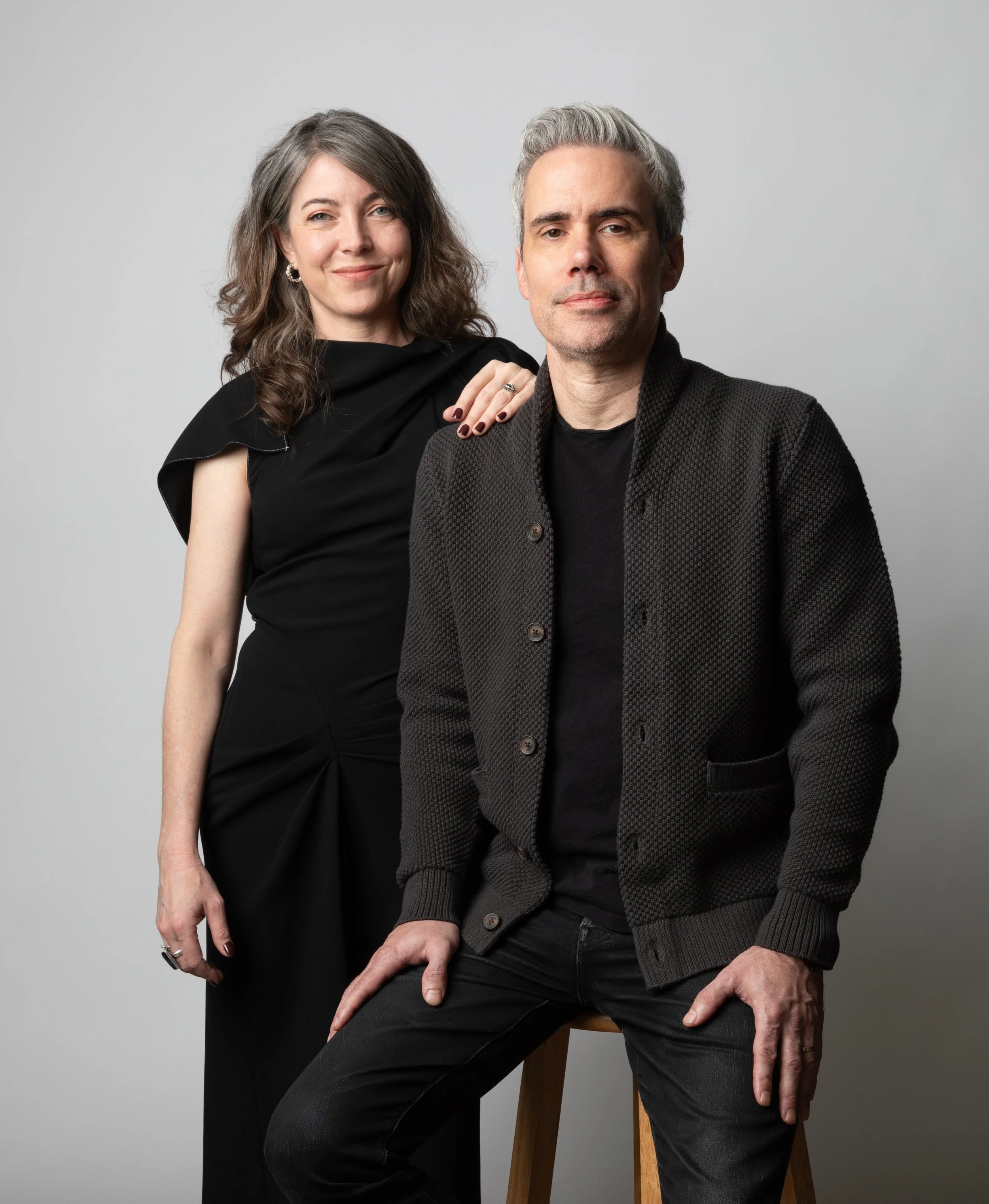 A woman with dark, wavy hair wearing a black dress with a shoulder detail, and a man with gray hair wearing a black textured jacket and black jeans, posing together against a plain gray background.