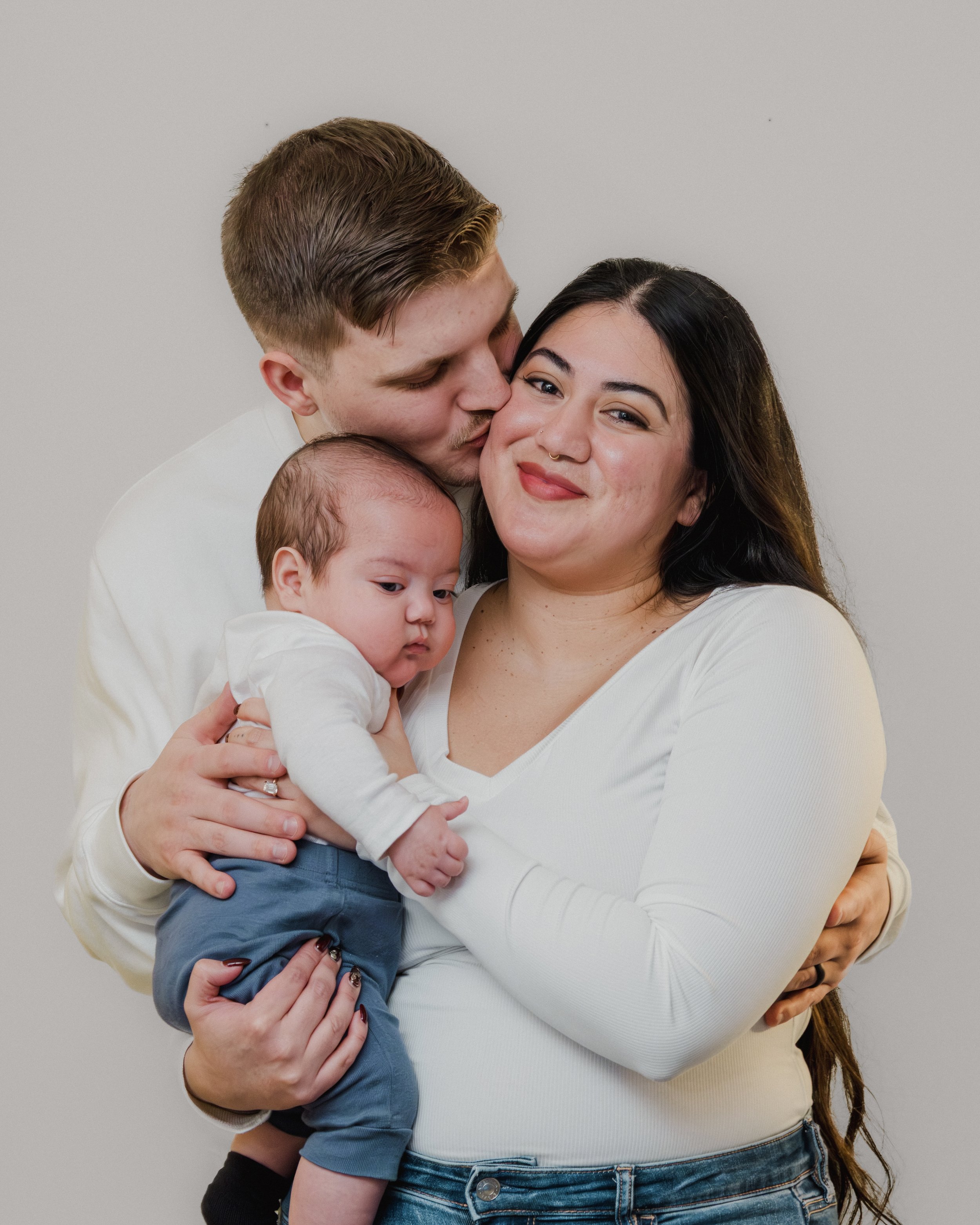 A family of three, including a woman holding a baby and a man kissing her on the cheek, posed against a plain background.
