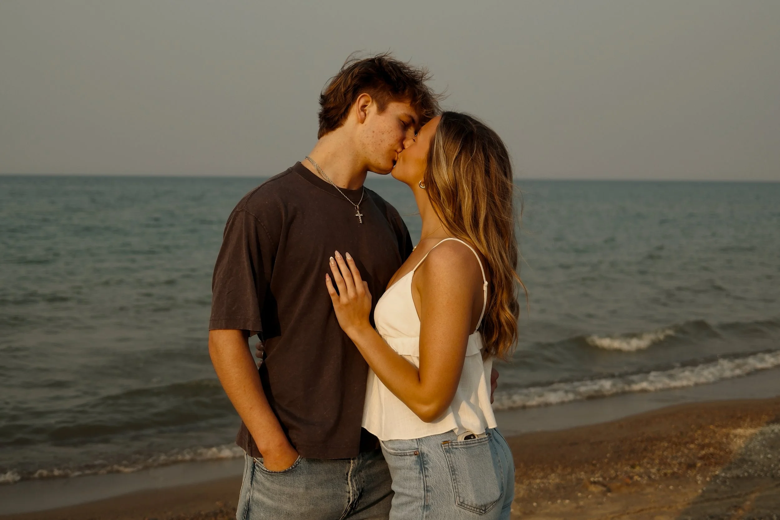 A young couple kissing on the beach during sunset, with ocean waves behind them.