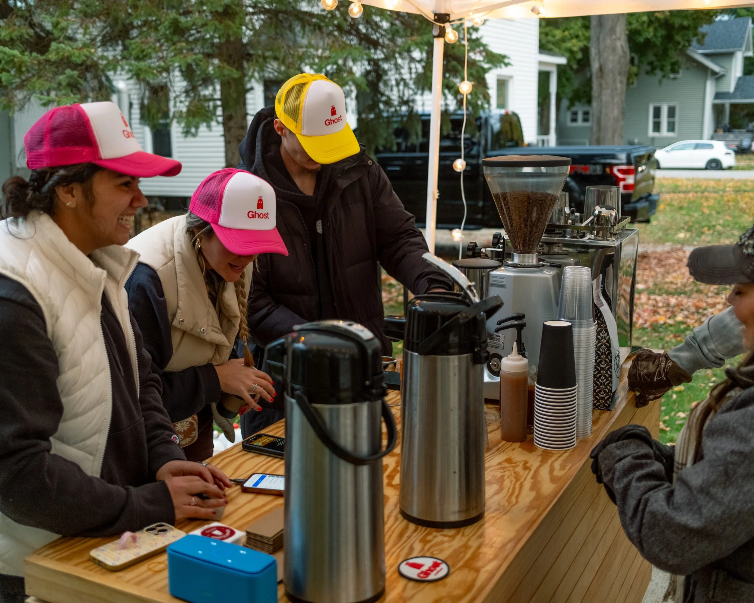 People at a coffee stand outdoors, wearing hats with the Ghost logo, preparing and ordering coffee.