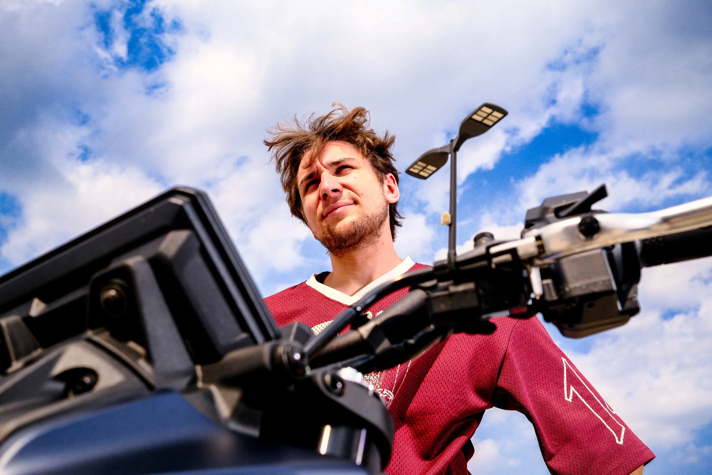 A man in a red sports jersey is looking up with a confused or perplexed expression while standing beside a motorcycle against a partly cloudy blue sky.