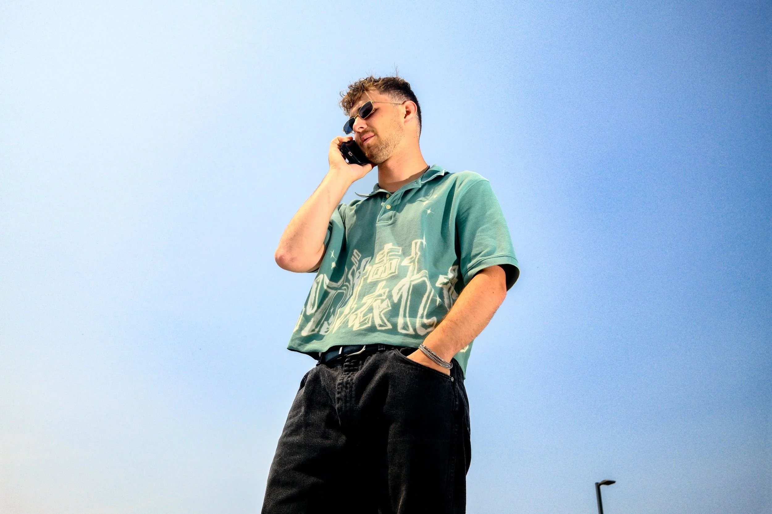 A young man with sunglasses, short curly hair, a green patterned polo shirt, and black pants, talking on a cell phone against a clear blue sky.