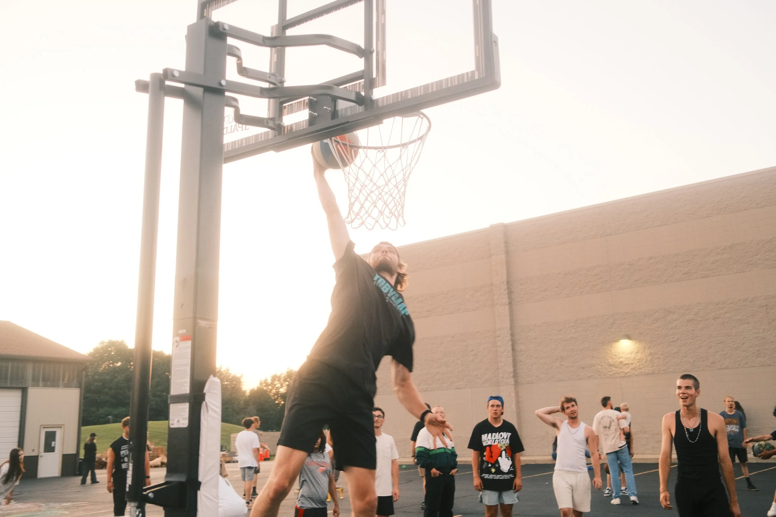 A man playing basketball outdoors, jumping to dunk the ball into the hoop, surrounded by onlookers at sunset.