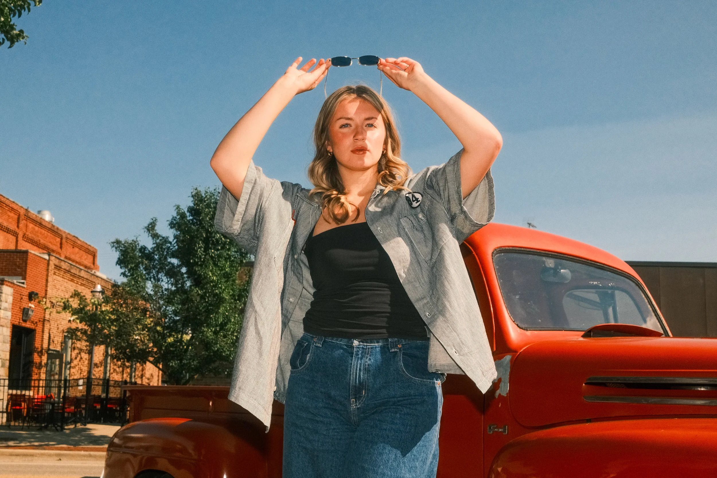 A young woman standing in front of a red vintage truck, holding sunglasses above her head, outdoors on a sunny day.