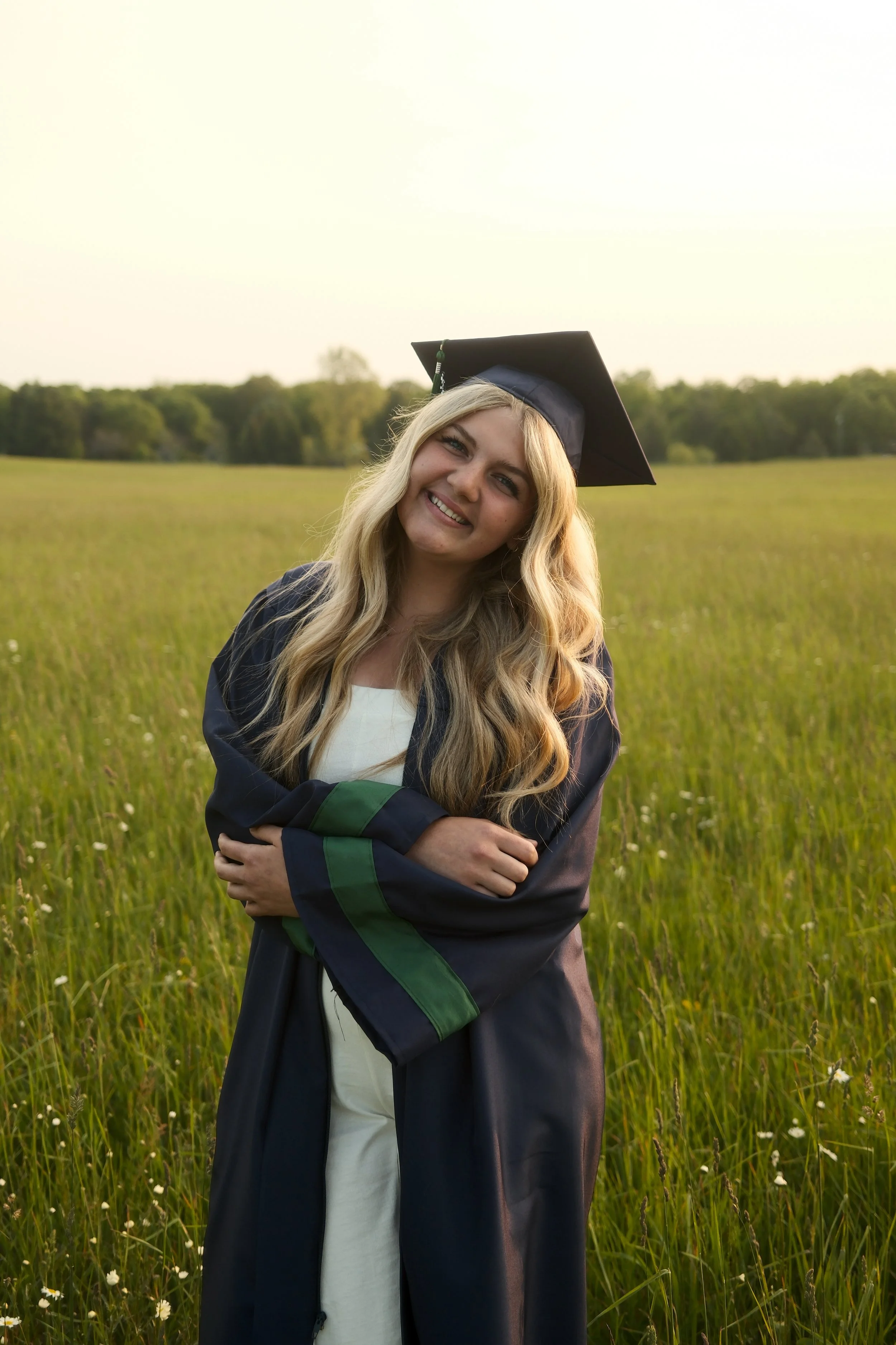 A young woman in a graduation cap and gown standing in a grassy field, smiling at the camera.