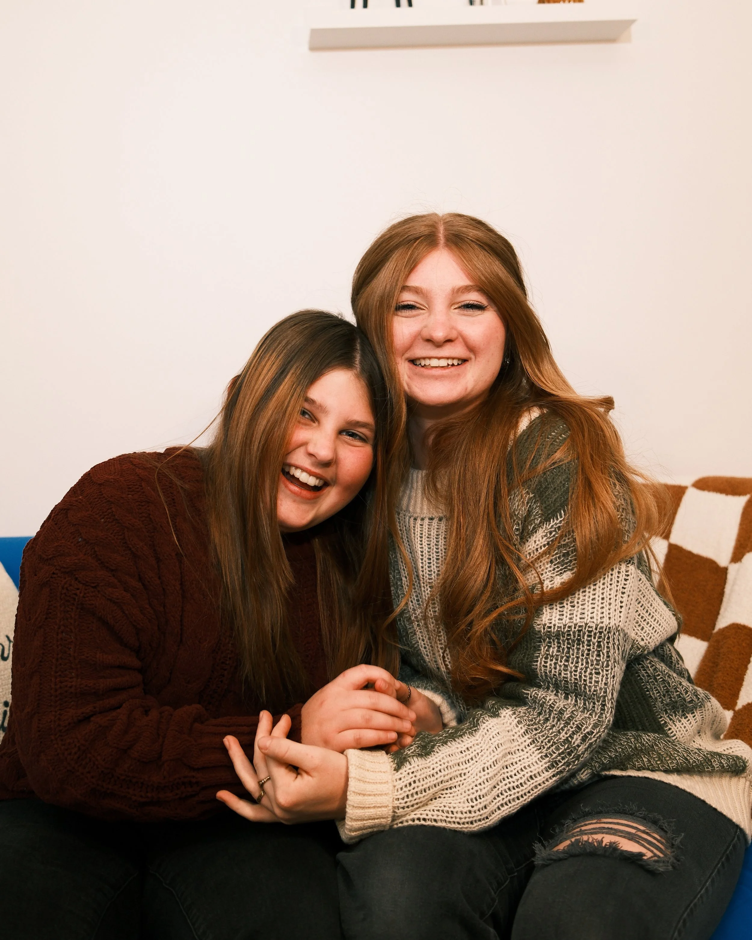 Two young women with long hair smiling and hugging each other on a couch in a cozy room.