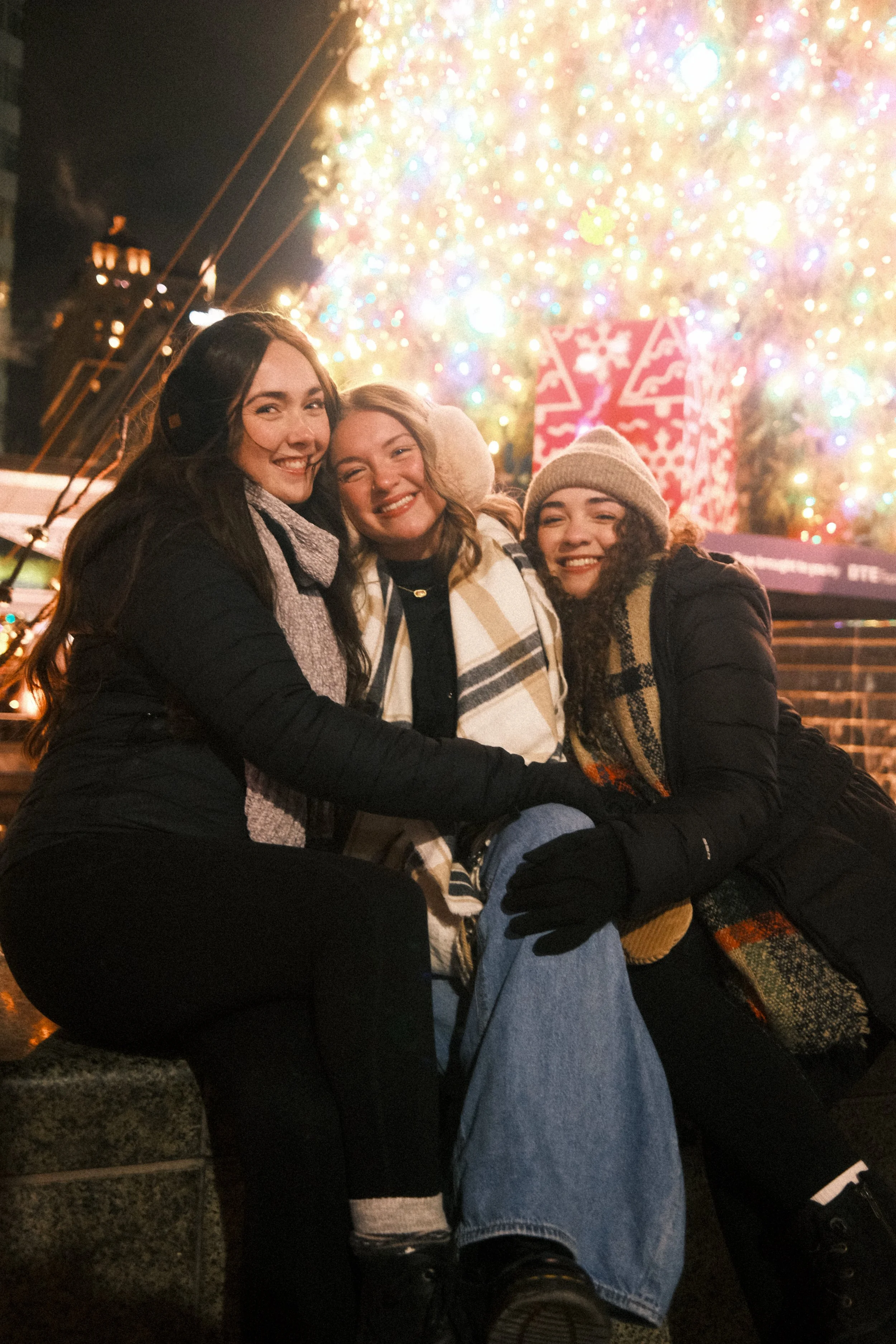 Three women dressed warmly, sitting together outdoors at night with a brightly lit Christmas tree and decorated gift box in the background.