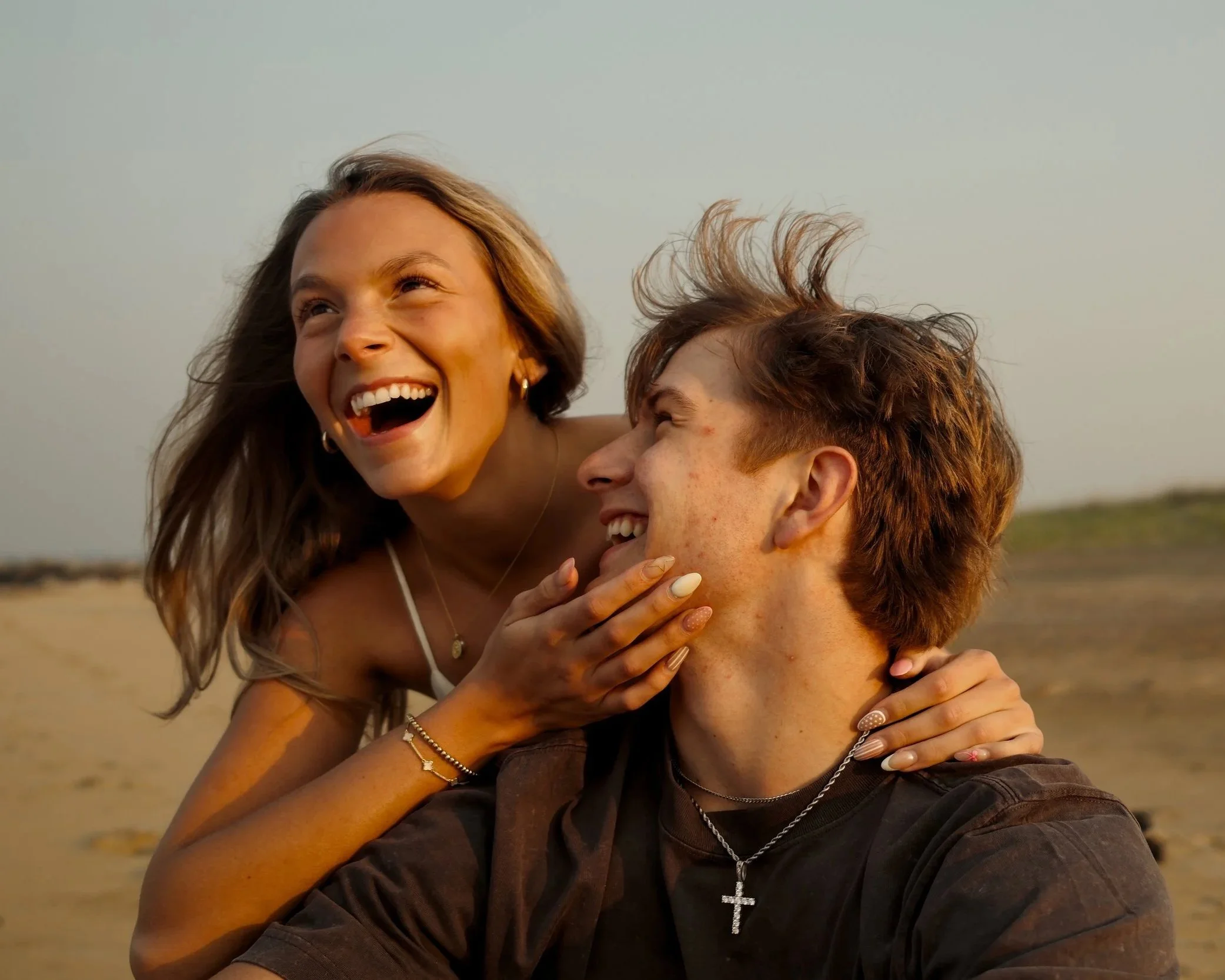 A young woman and man sharing a joyful moment on the beach, smiling and laughing with the woman touching the man's face.