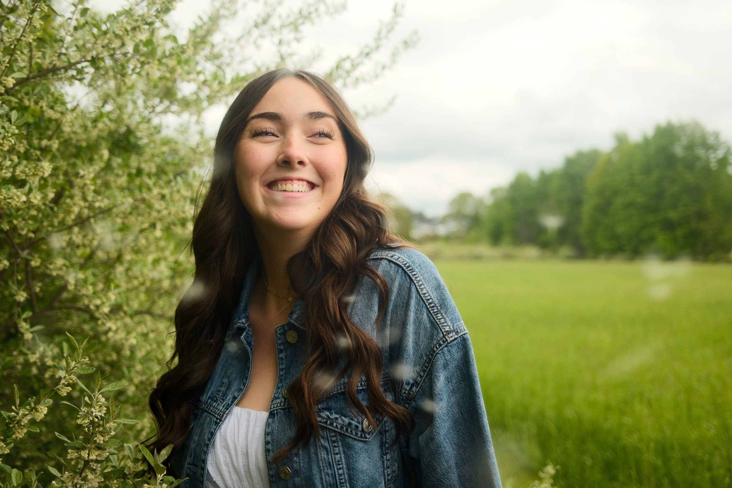 A young woman with long brown hair smiling outdoors next to green bushes and a field.