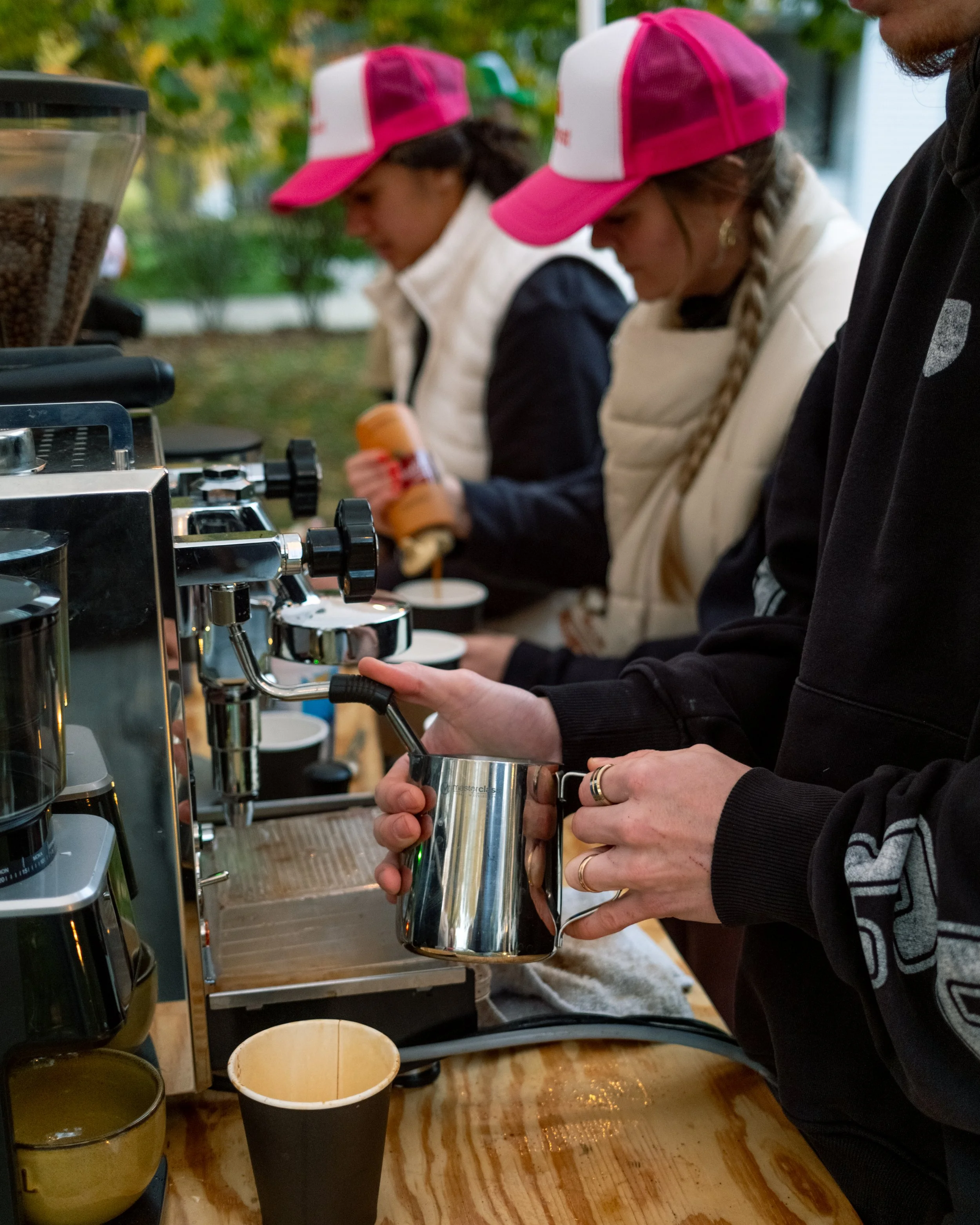 People making coffee at an outdoor coffee stand, with three women in pink and white hats and vests, and a person holding a milk frothing pitcher.