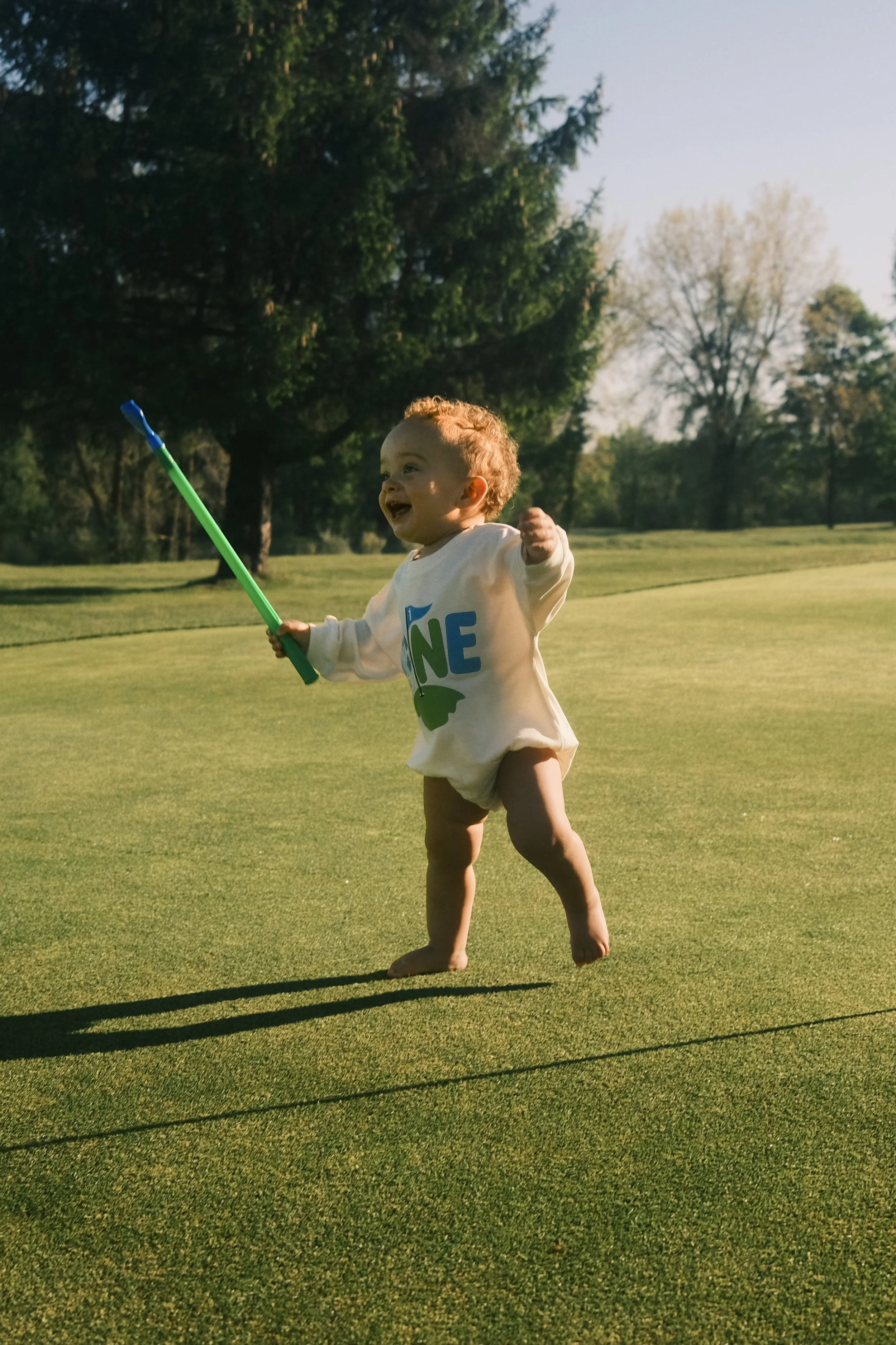 A baby standing on a grassy field, holding a small golf club, smiling and pointing with joy, with trees and a clear sky in the background.