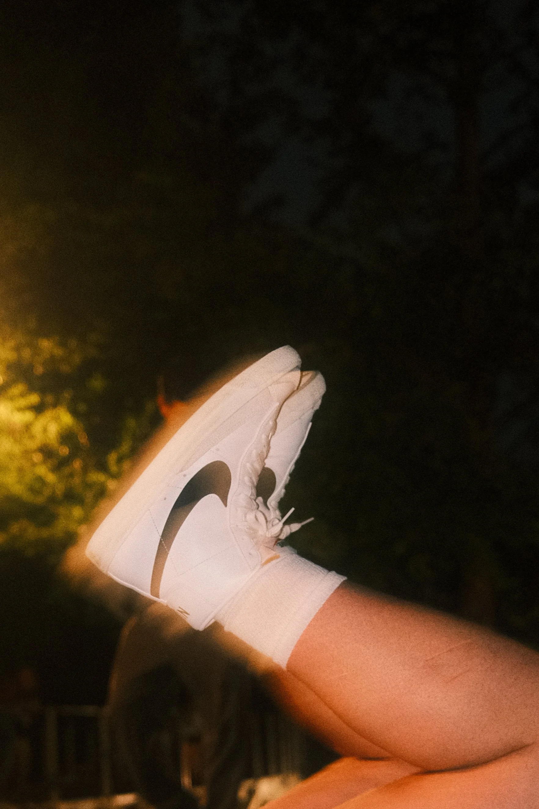 Close-up of a person's leg in a white Nike sneaker with a black swoosh logo, illuminated by a yellowish light at night, with a dark background and some trees.