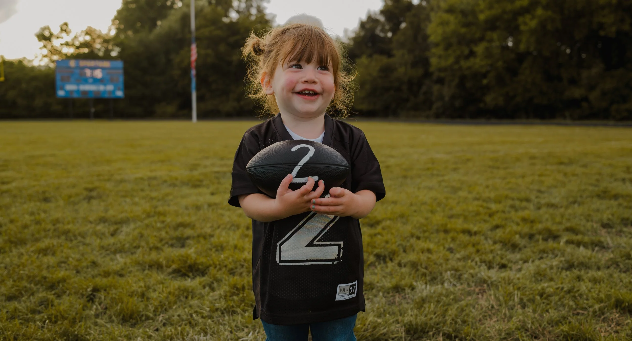 A young girl with brown hair in pigtails, wearing a black football jersey, holding a black football with a white question mark on it, standing on a football field during sunset with trees in the background.