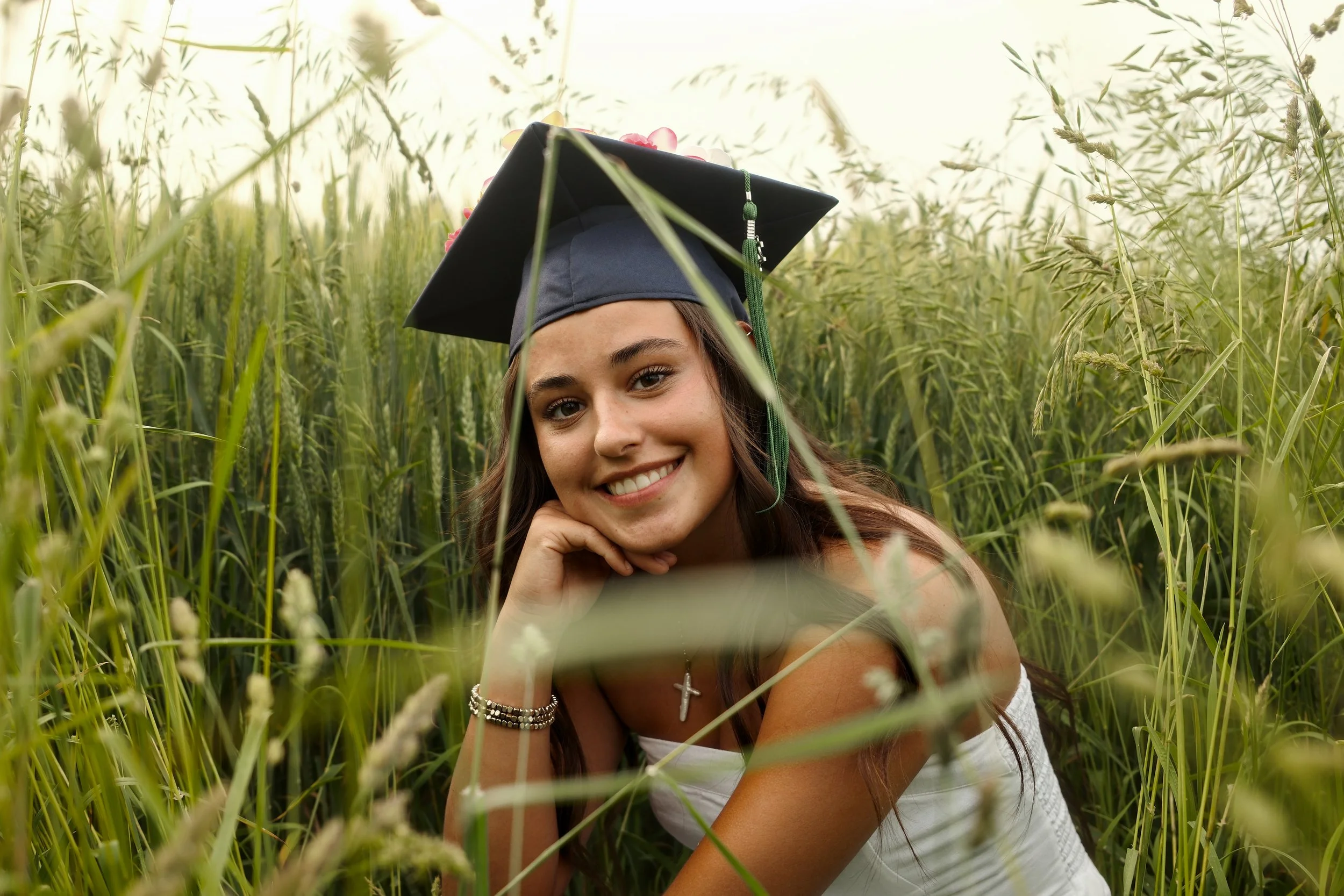 Young woman with long brown hair, wearing a graduation cap and white dress, lying in a field of tall green grass, smiling at the camera. Senior photos, cap and gown photographer in Michigan.