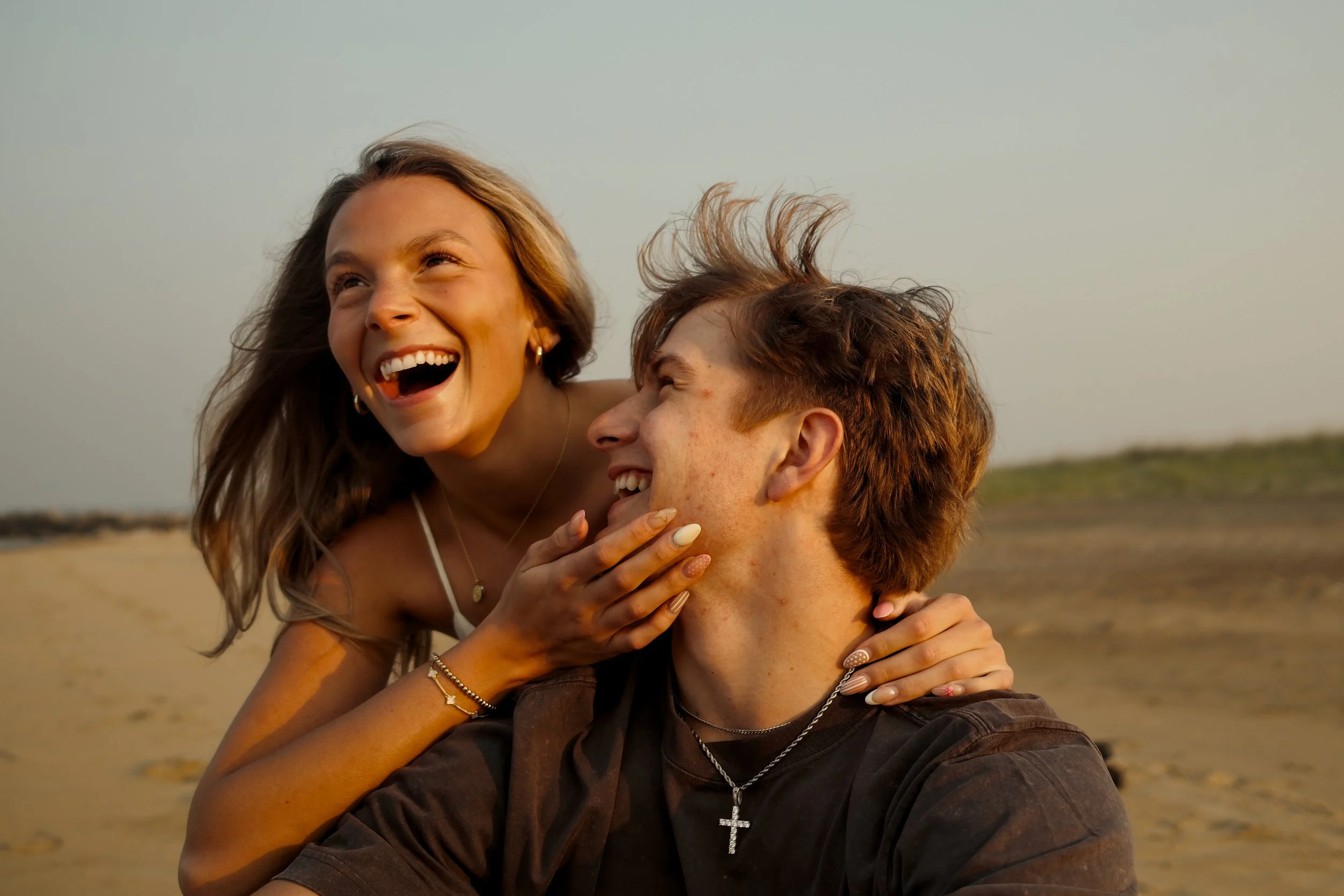A happy young couple at the beach, with the woman playfully touching the man's chin, both smiling and enjoying each other's company.