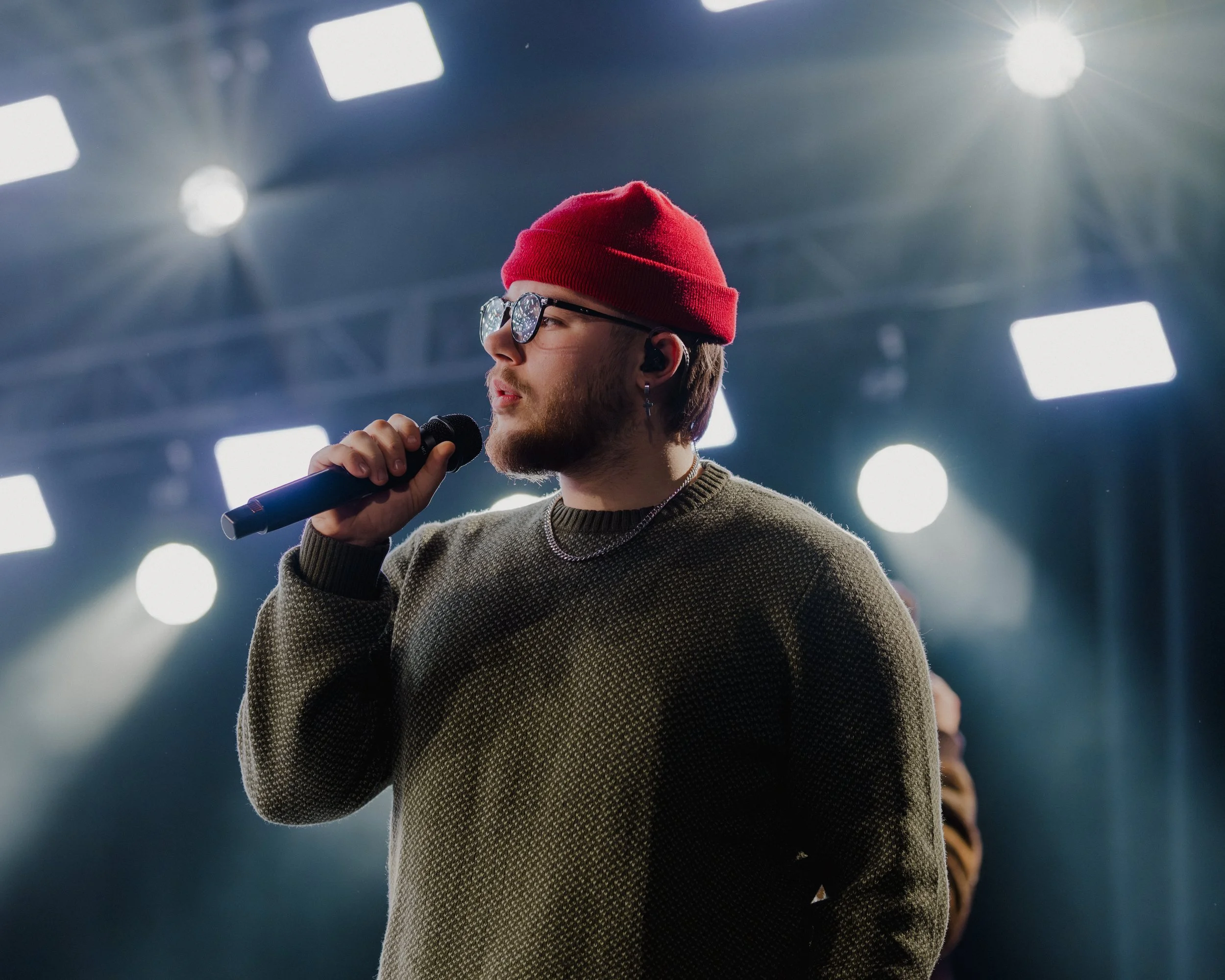 A young man with glasses, a red beanie, and a sweater, holding a microphone and singing or speaking on stage with bright lights in the background.