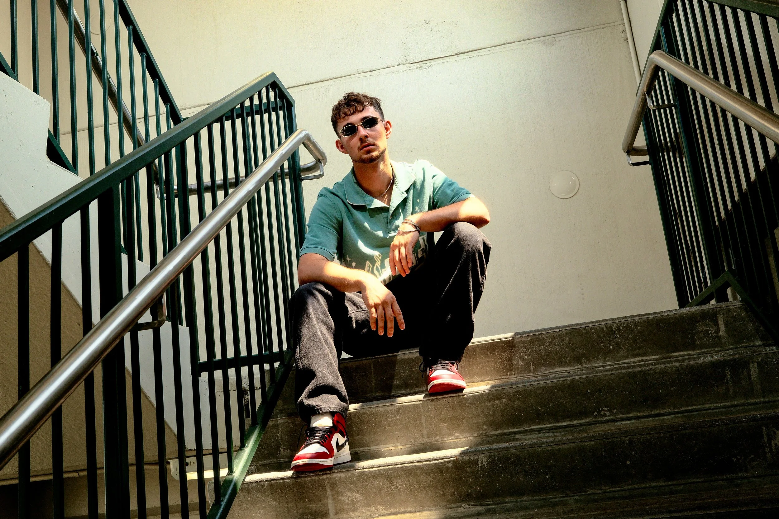 Young man sitting on stairs between green metal railings, wearing sunglasses, a green shirt, black pants, and red, white, and black sneakers.