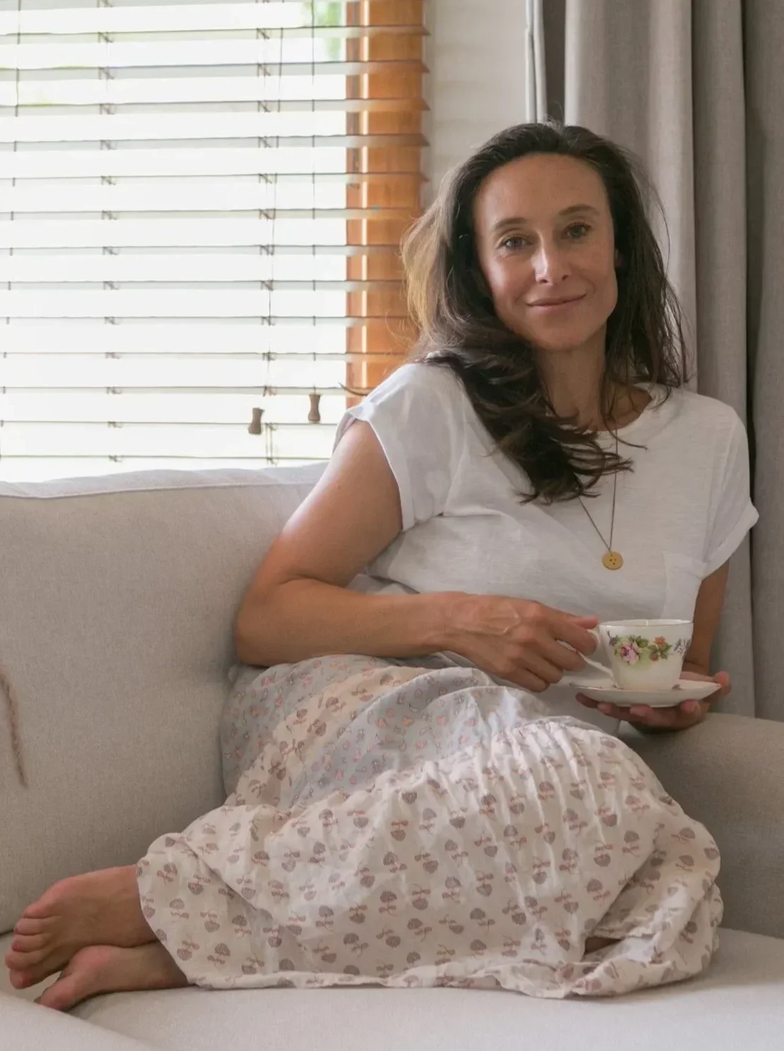 A woman sitting on a sofa, holding a teacup and saucer, wearing a white t-shirt and patterned pants, with brown hair, in a room with window blinds and curtains.
