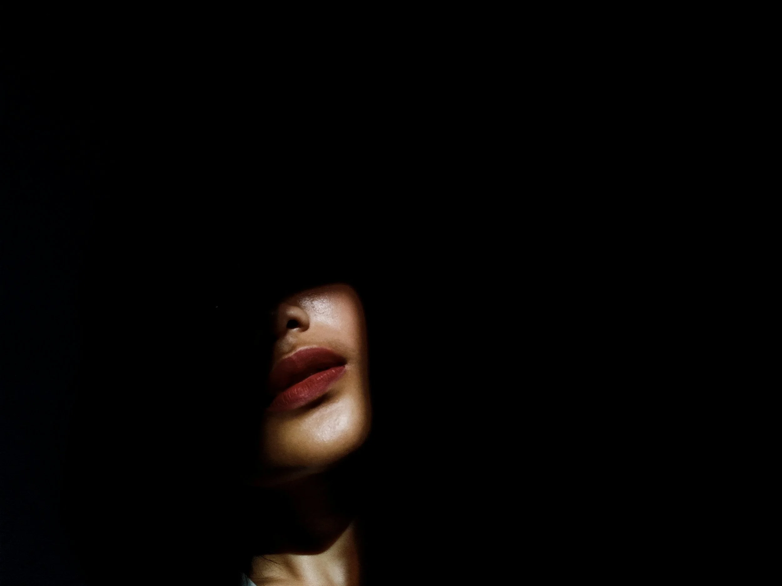 Close-up of a woman's face partially covered in darkness, showing lips and a nose with soft lighting.
