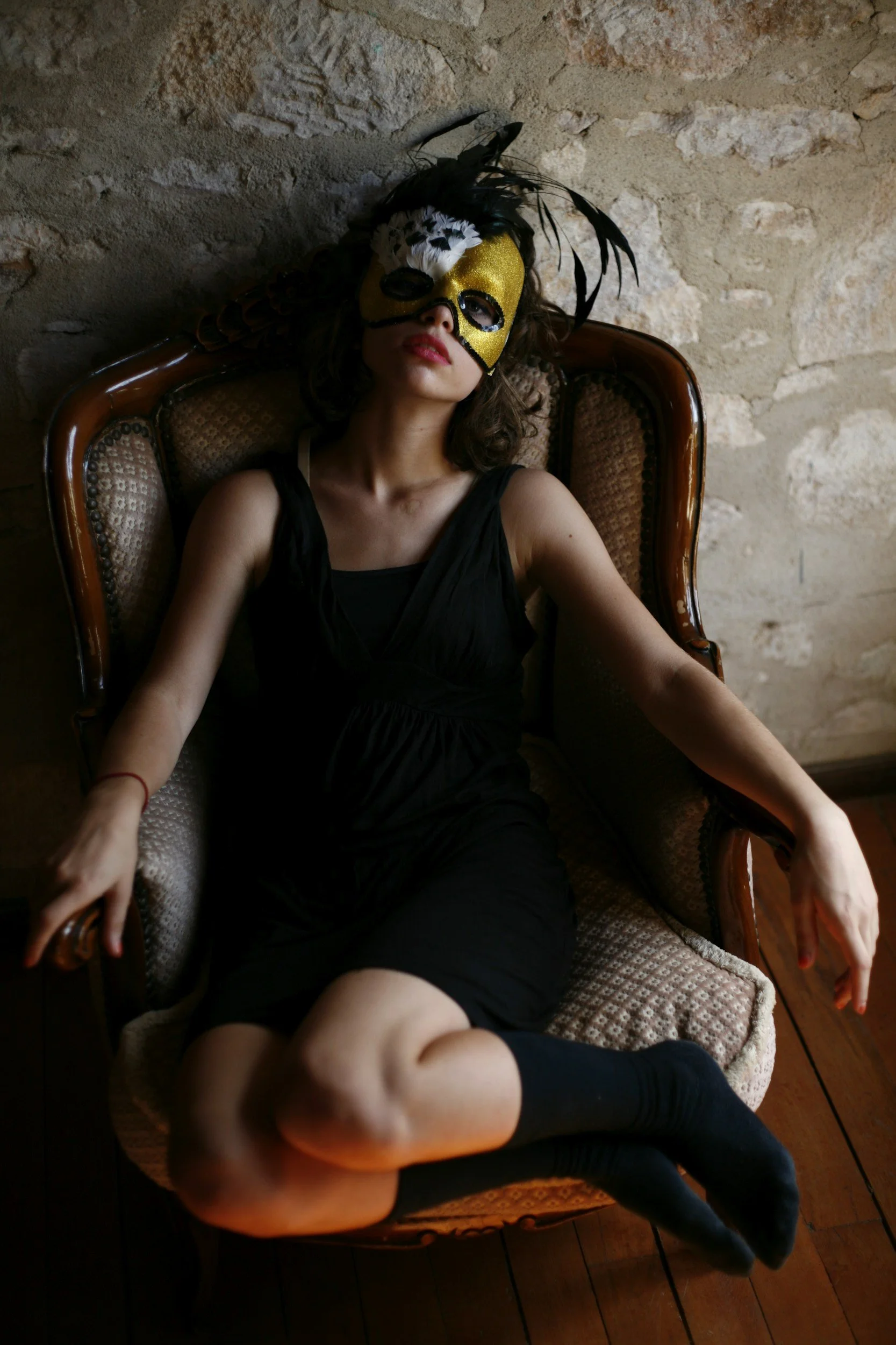 A woman is sitting on a vintage armchair with a stone wall behind her. She is wearing a black dress, black socks, and a gold and black mask with feathers covering her face.