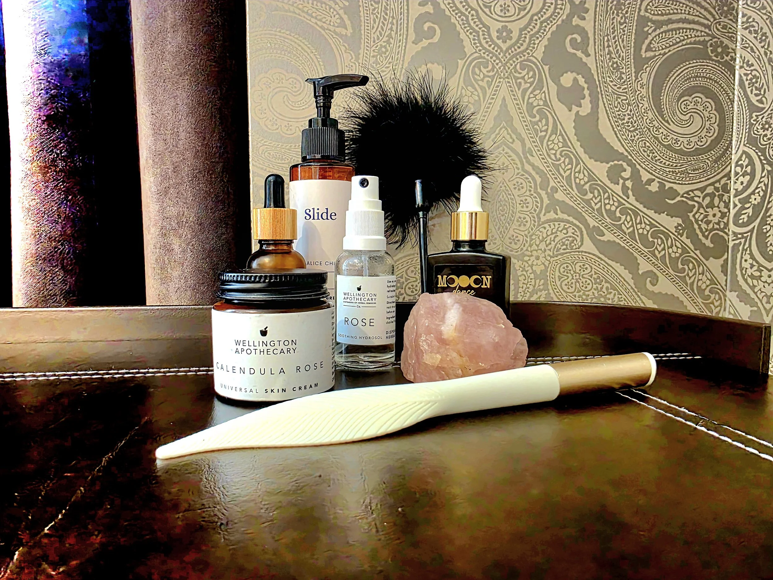 A collection of skincare and beauty products on a wooden surface, including a jar labeled 'Wellington Apothecary Calendula Rose Universal Skin Cream,' a bottle of rose soothing hydrosol, a glass dropper, an amber bottle labeled 'Slide,' a black bottle labeled 'Moon,' a pink salt crystal, a black fluffy brush, and a beige and white facial roller, with a decorative background and a vertical wooden object.