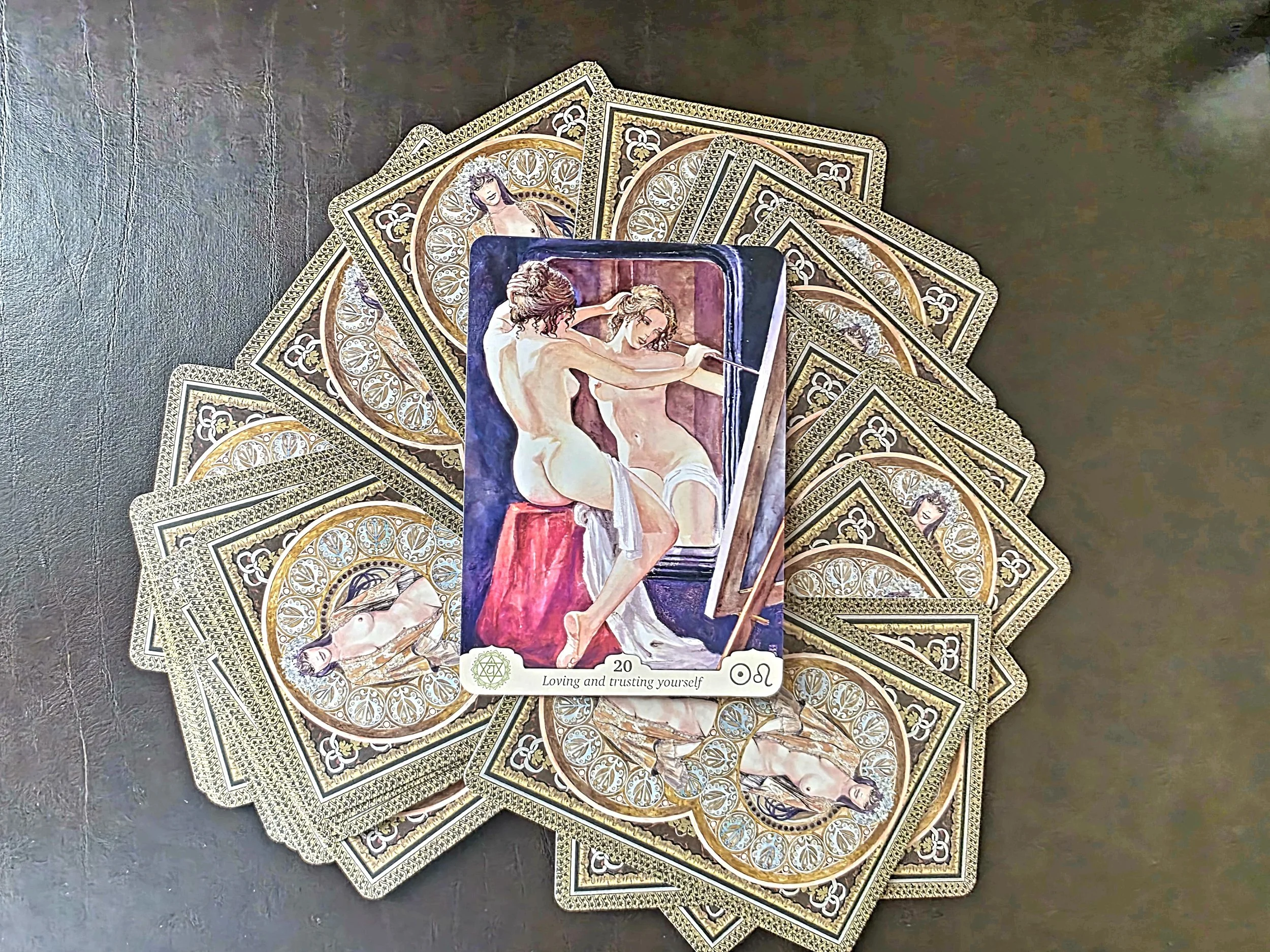 A tarot card reading spread with a detailed art card placed on top, depicting two women in an intimate pose with one holding a sword, and back drop of deep red and purple colors. The spread contains multiple tarot cards with ornate gold and silver designs, each featuring different nude or scantily clad women.