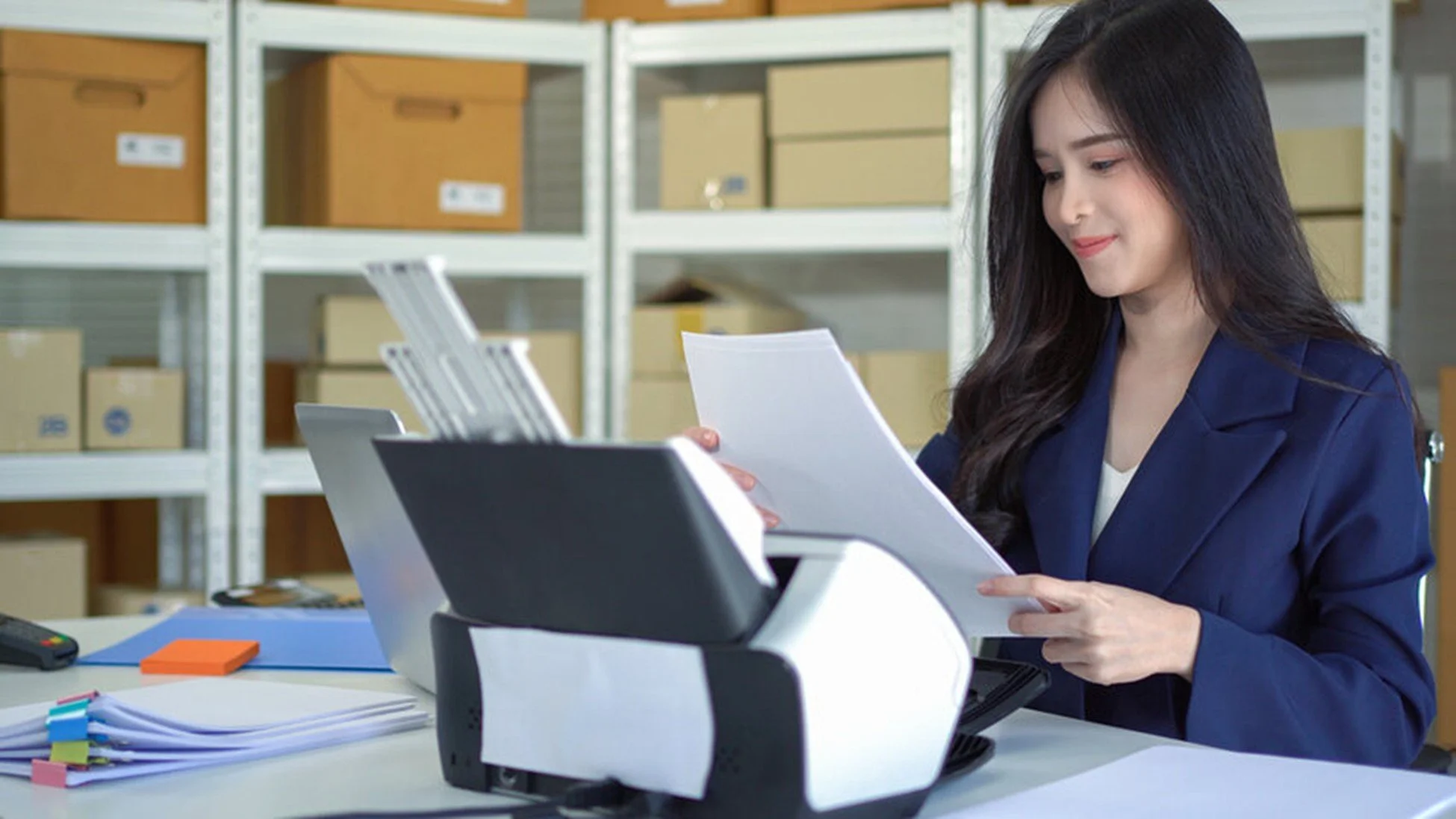 Woman working at office desk with printer and files, storage boxes in the background.