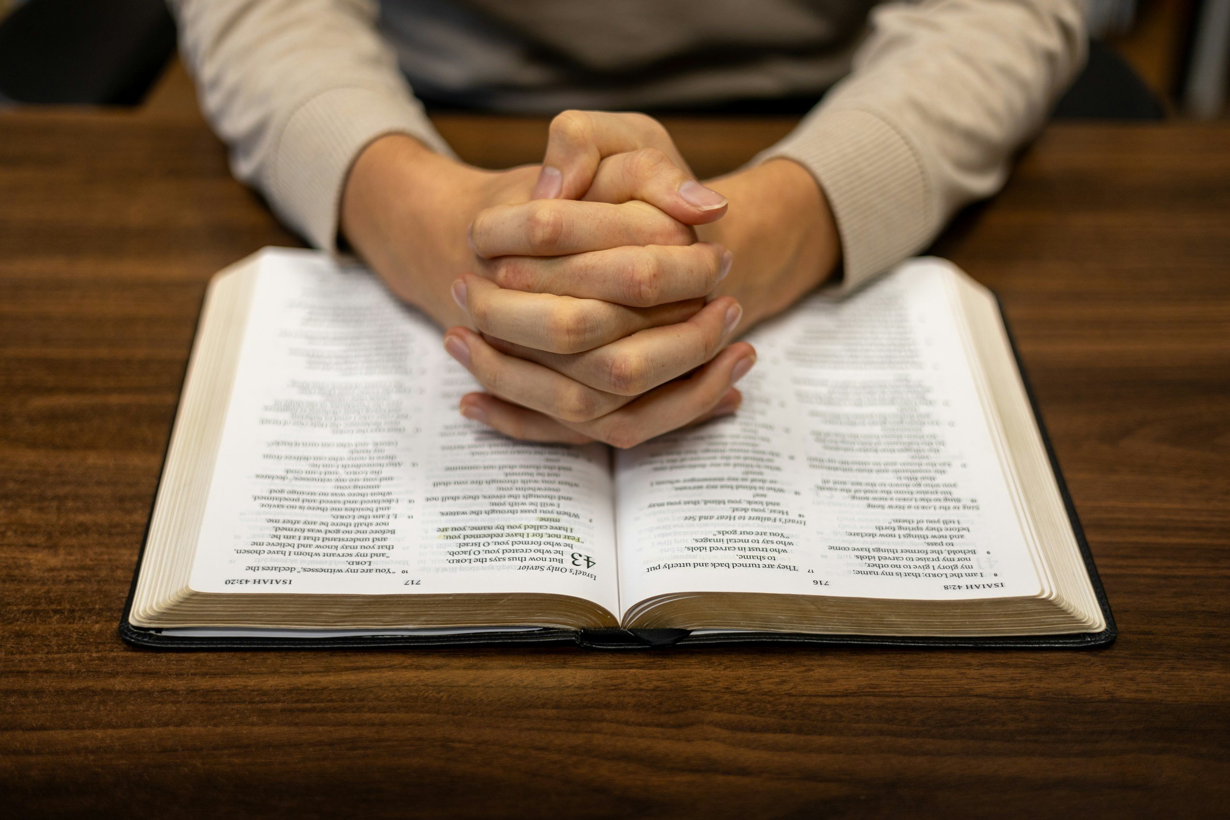 Person with hands clasped in prayer over an open Bible on a wooden table.
