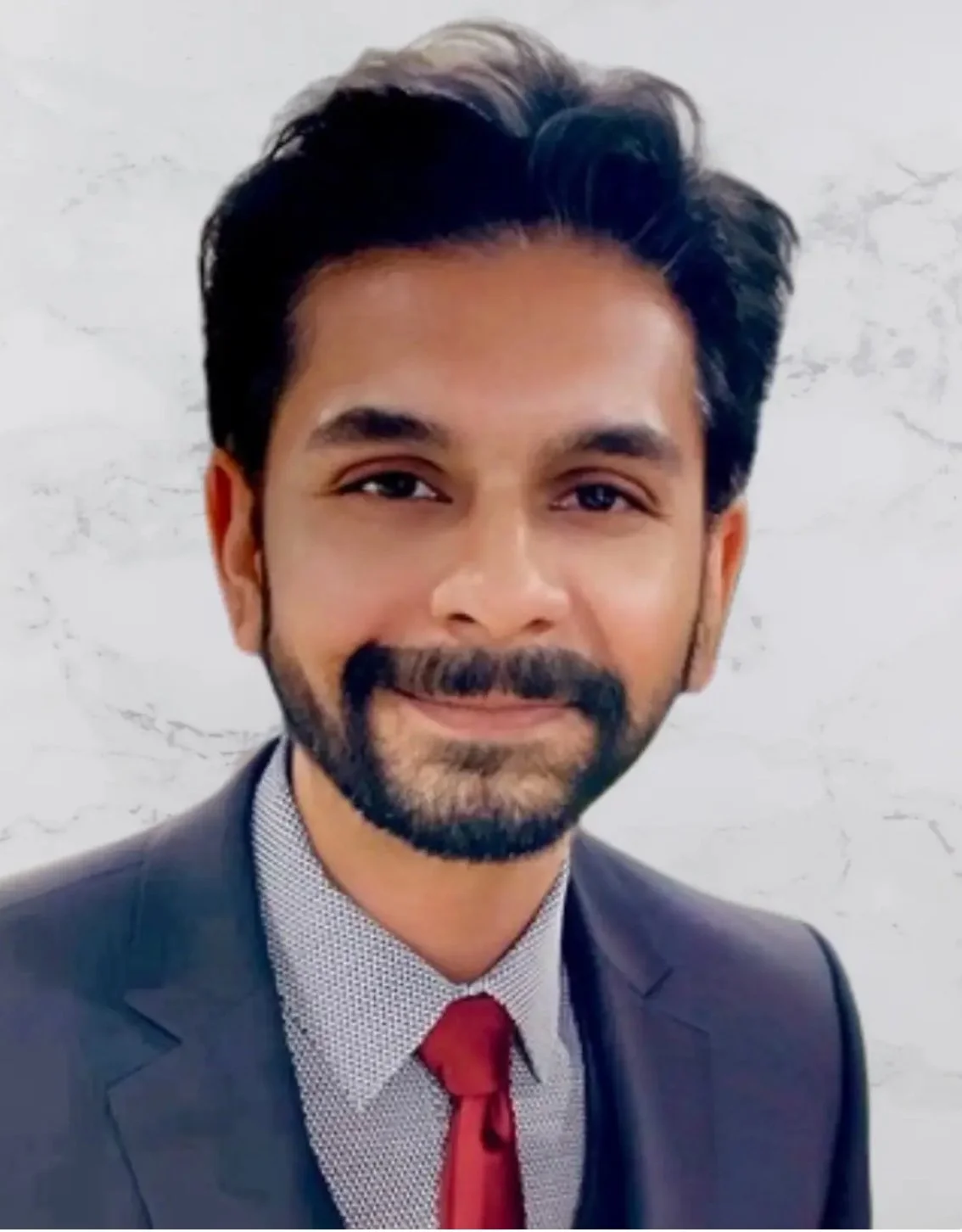 A man with dark hair and beard, wearing a suit, white shirt, and red tie, smiling in front of a white wall.