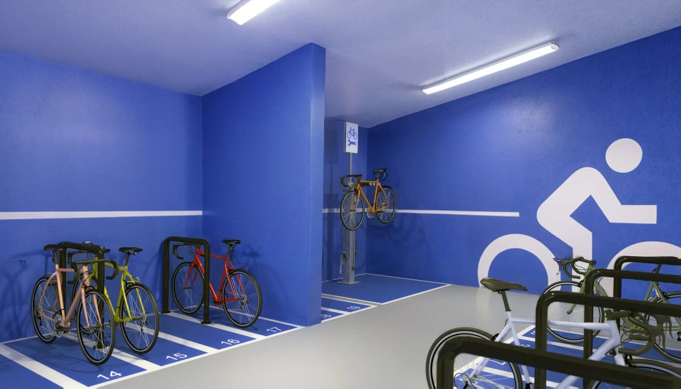 Indoor bike parking area with multiple bicycles stored in designated numbered racks, a large bicycle symbol painted on the wall, and a blue wall with a white line running horizontally across it.