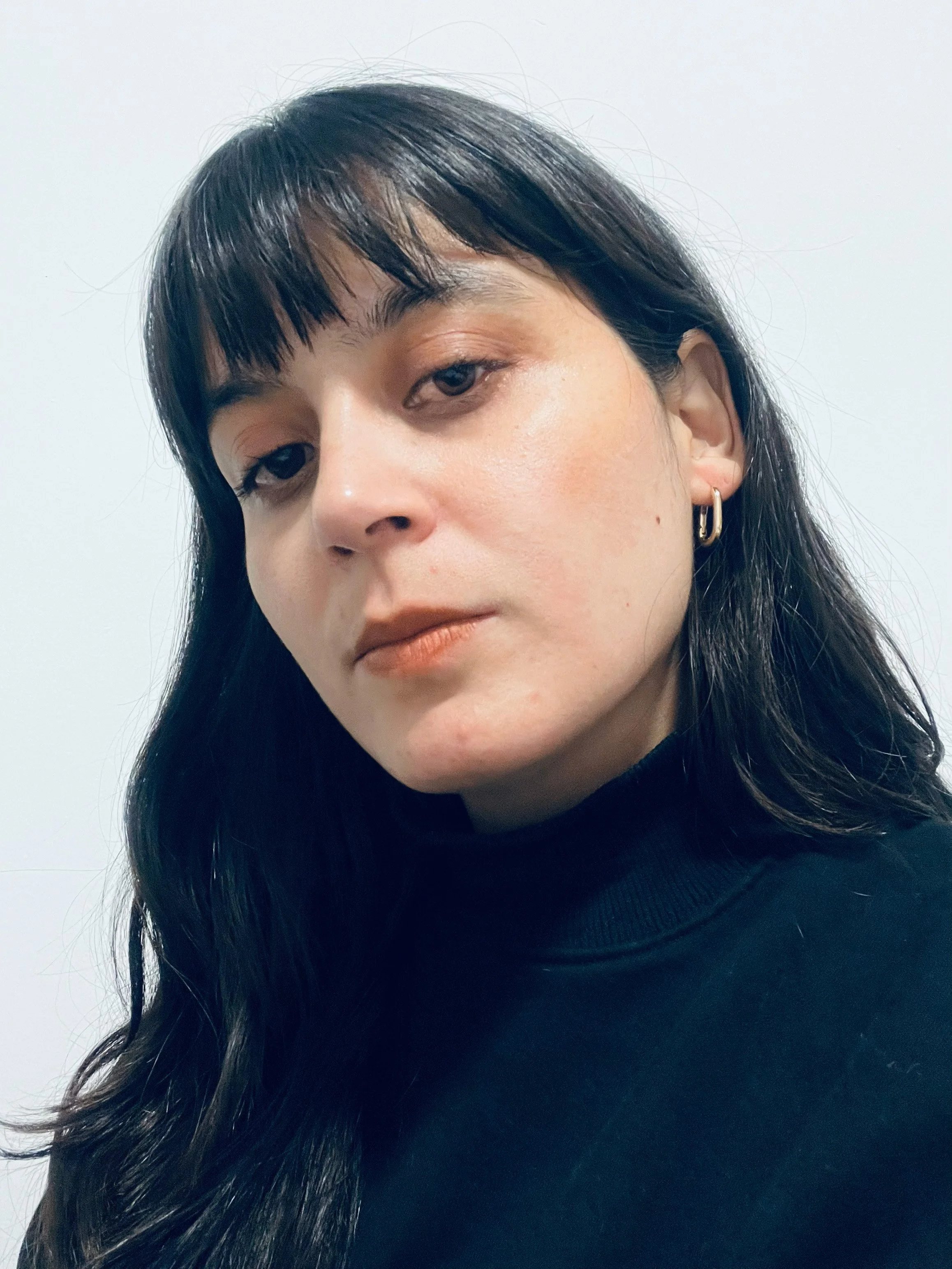Close-up of a young woman with dark hair, wearing a black turtleneck and gold hoop earrings, against a plain white wall.