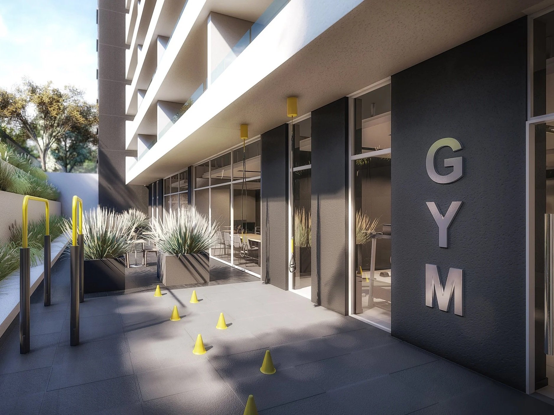 Modern building entrance with glass doors and windows, potted plants with tall grasses, yellow traffic cones and barriers, and a sign with large metal letters spelling GYM on a dark wall.