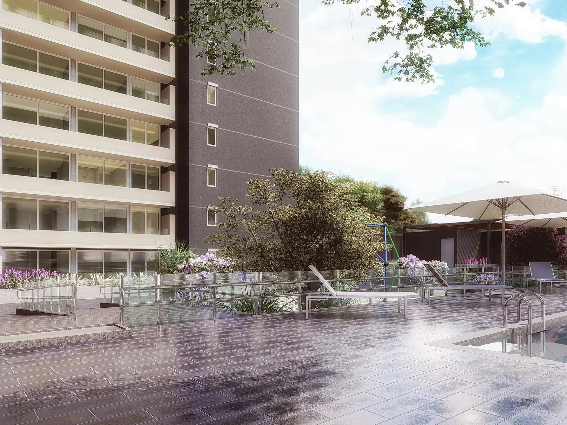 View of a modern apartment building with a patio area featuring lounge chairs, an umbrella, garden plants, and a rubber playground in the background, with trees and blue sky.