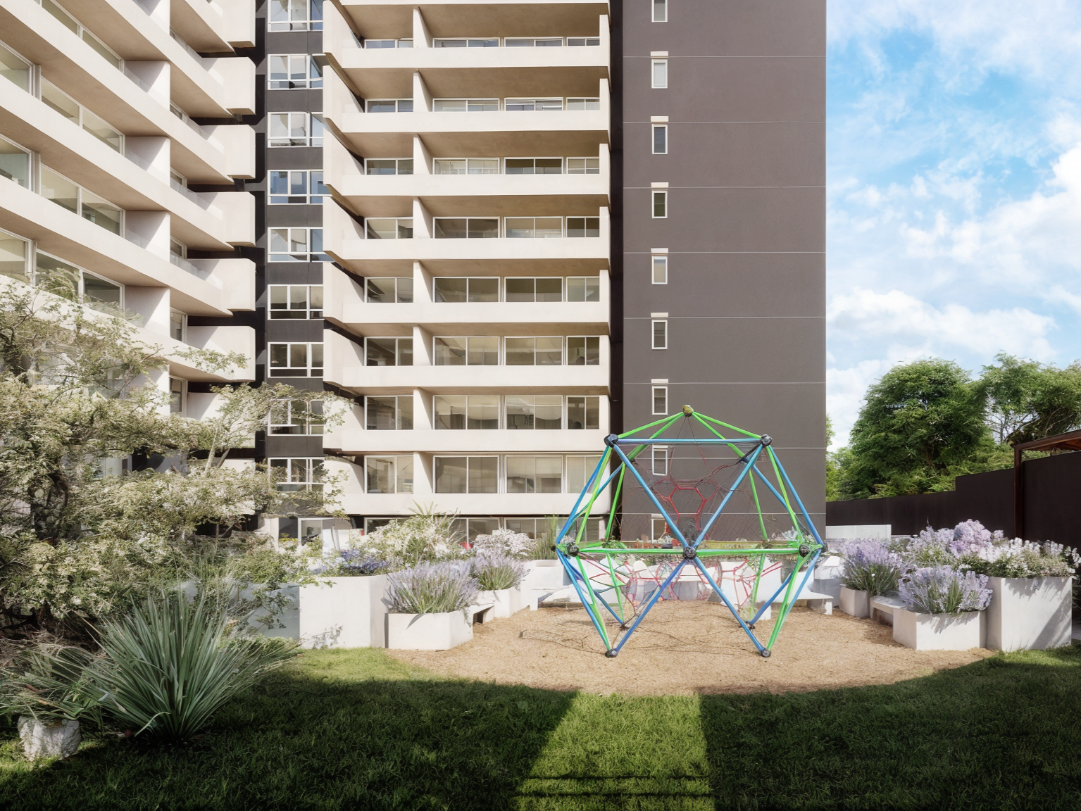 A colorful climbing structure in a small courtyard with lush plants and flowers, in front of a modern multi-story apartment building under a partly cloudy sky.