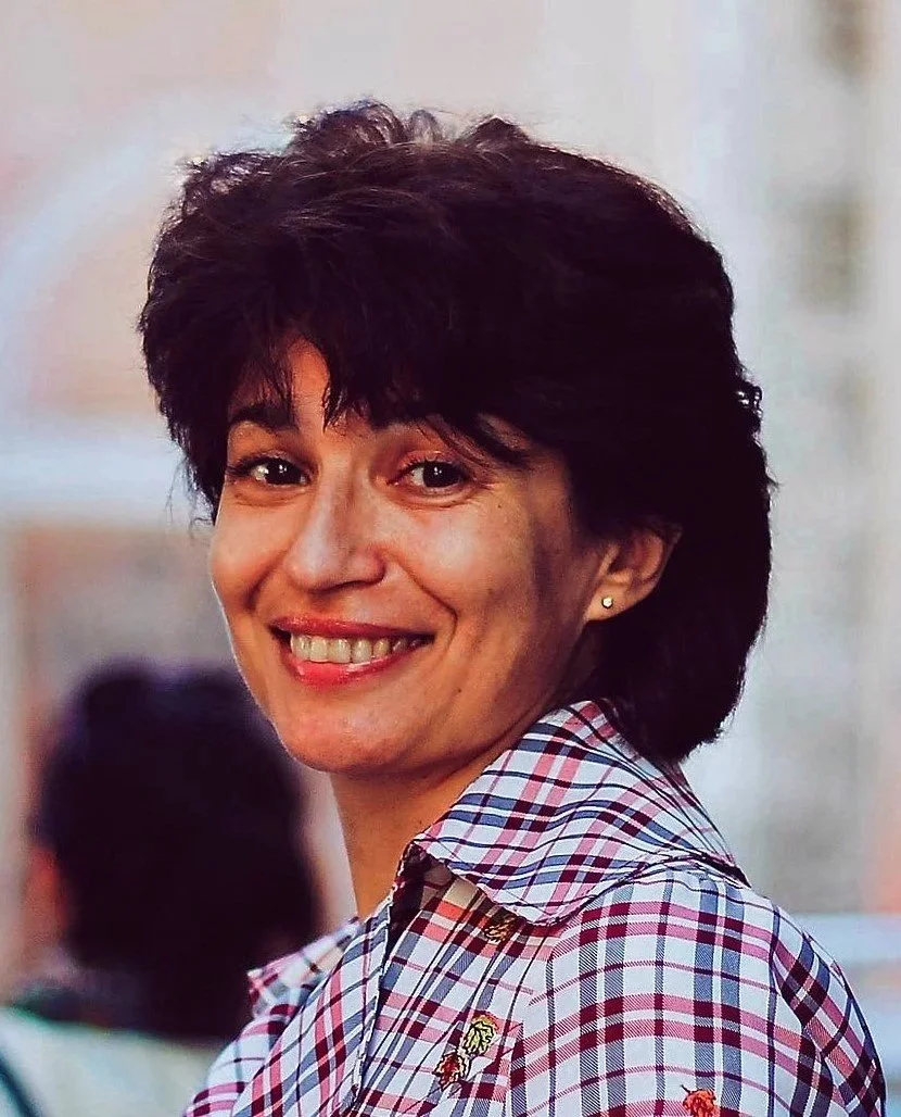 Illustrator Olena Kharkovenko, who has short dark hair, is  smiling,  and is wearing a checkered shirt with small embroidered animal patches, in an outdoor setting.