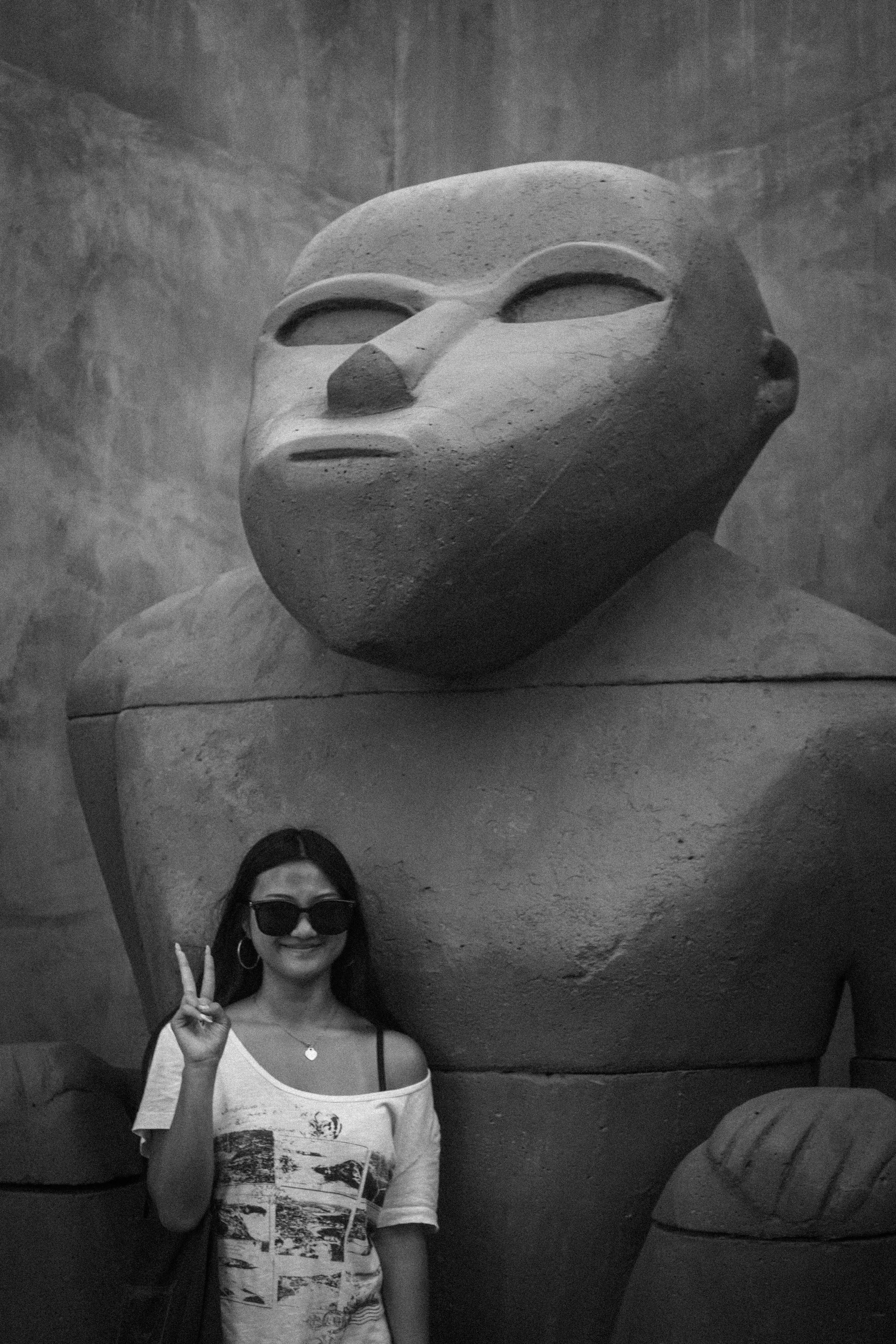 Woman wearing sunglasses poses with a peace sign beside a large stone sculpture in a museum.