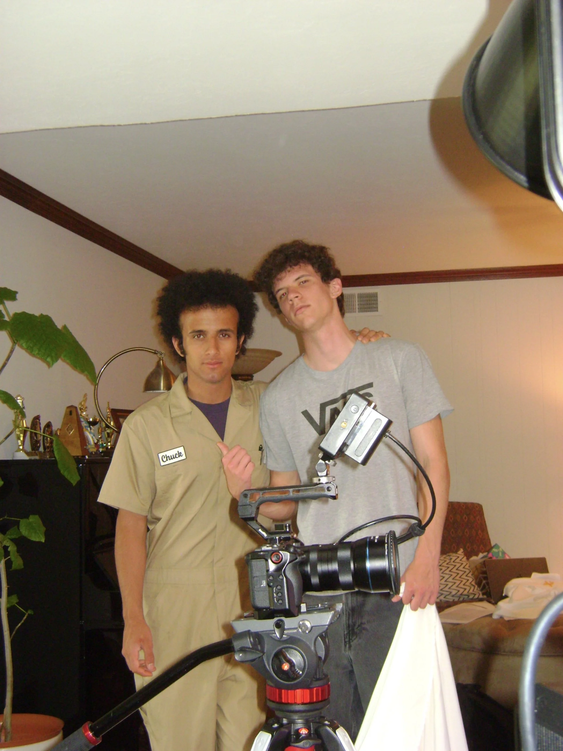 Two young men pose indoors beside a professional video camera on a tripod.