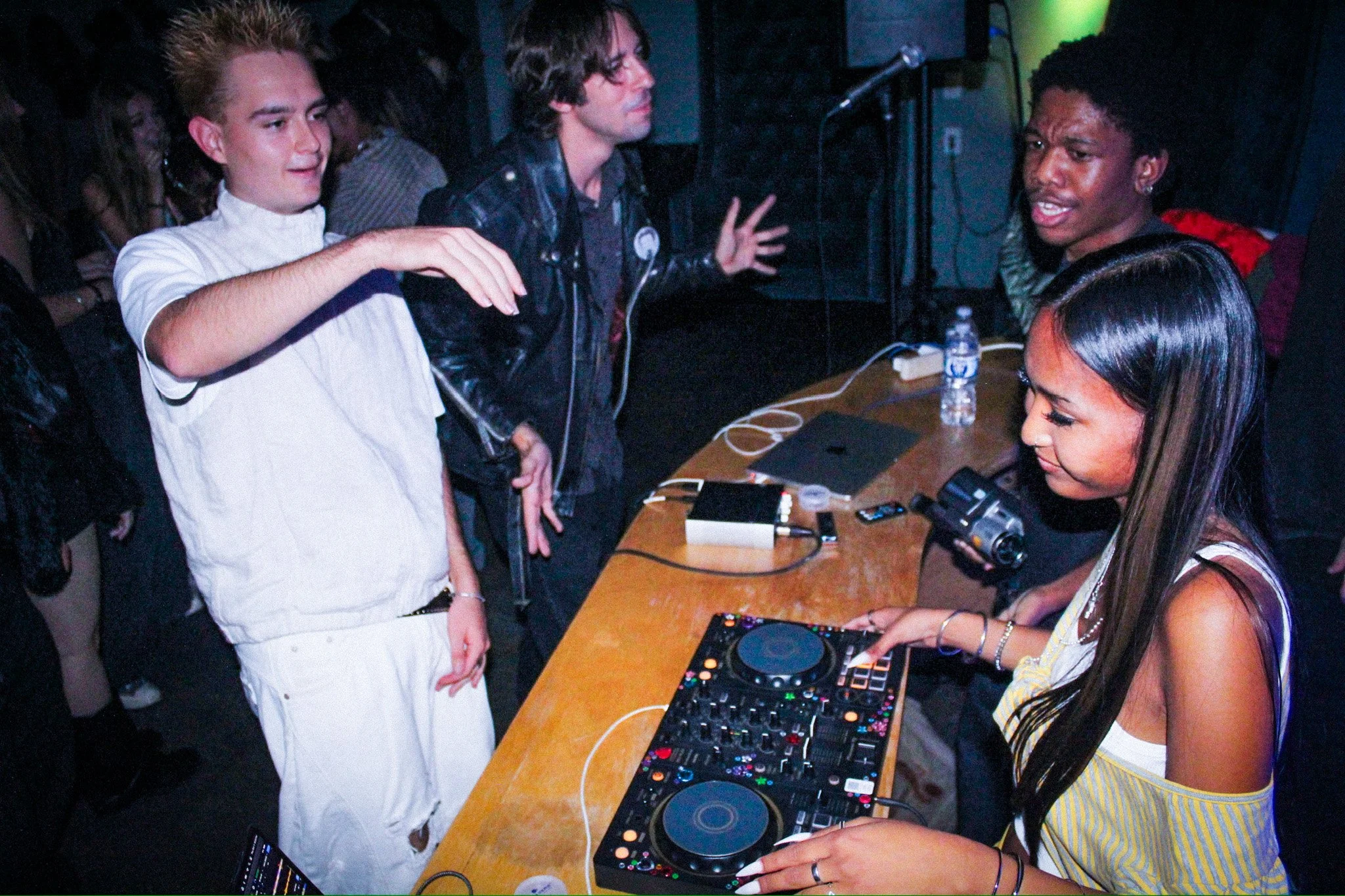 DJ operating a mixing console at a crowded nightclub party with people dancing nearby