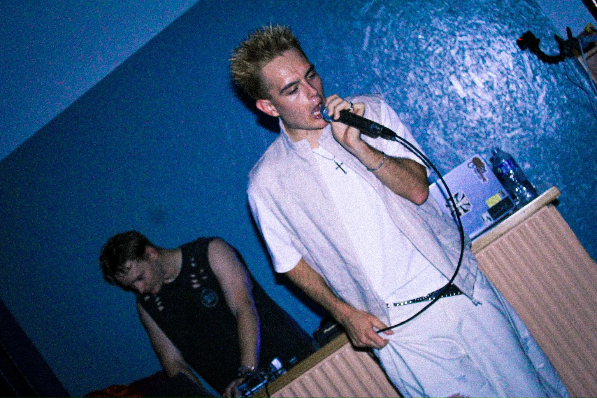 Male singer performing into a microphone onstage beside a guitarist in a dim blue-lit venue