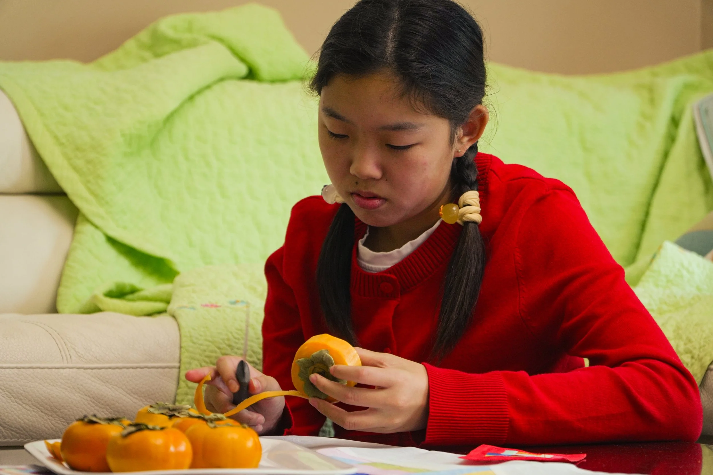 Young woman in a red sweater peeling a persimmon beside a plate of fruit.