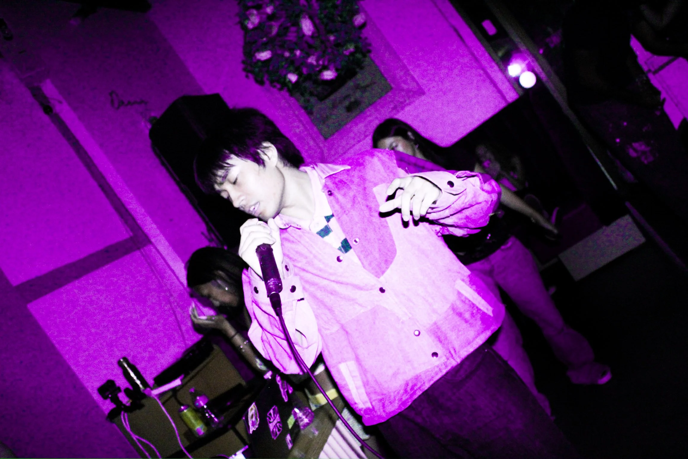 Singer performing into a microphone onstage under vivid purple lighting, with band members and equipment in the background.