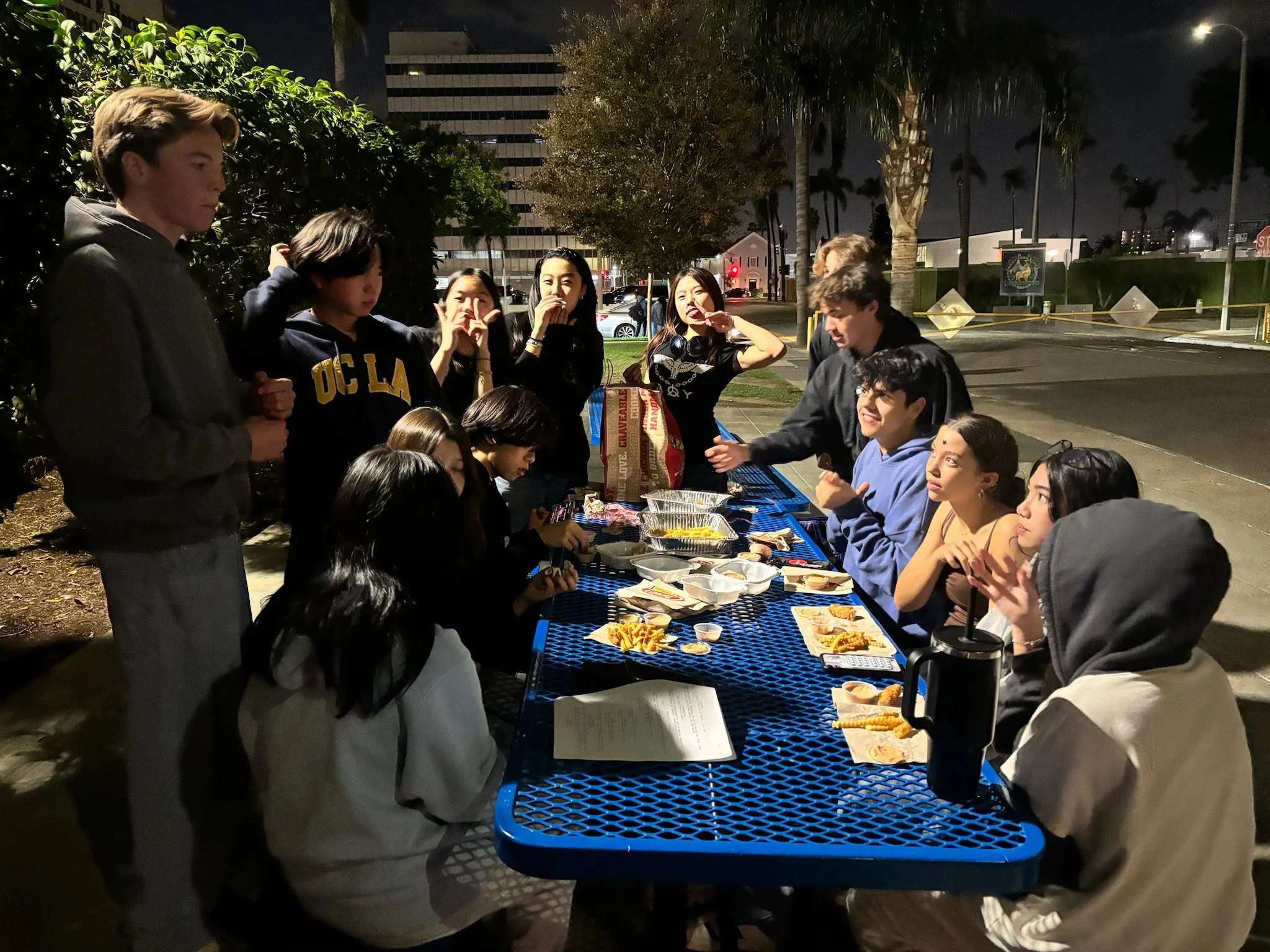 Group of young people eating takeout at a blue picnic table outdoors at night.