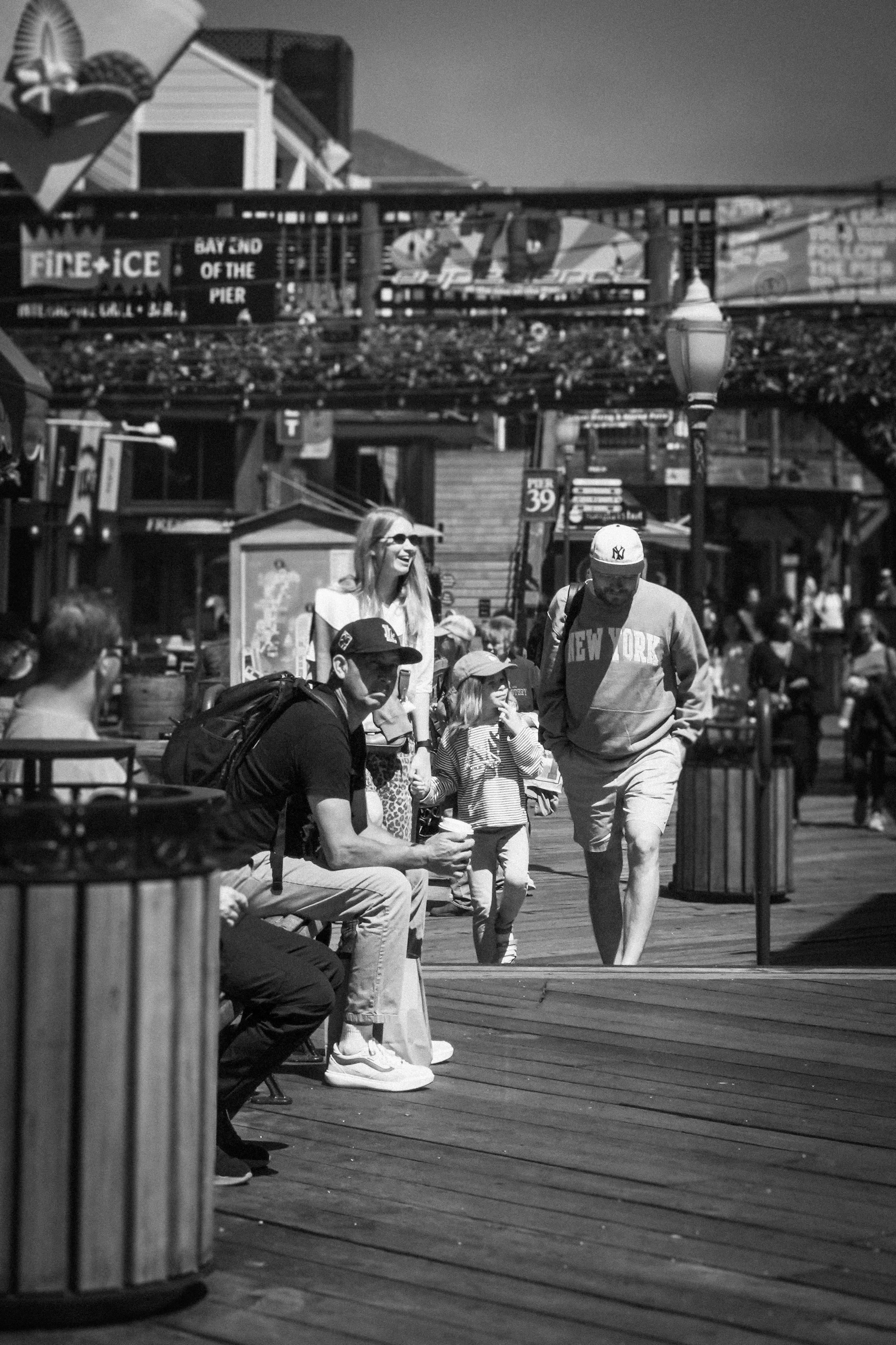 Black-and-white photo of tourists walking on a crowded pier beneath a roller coaster, with waterfront amusement park signs and rides in the background.