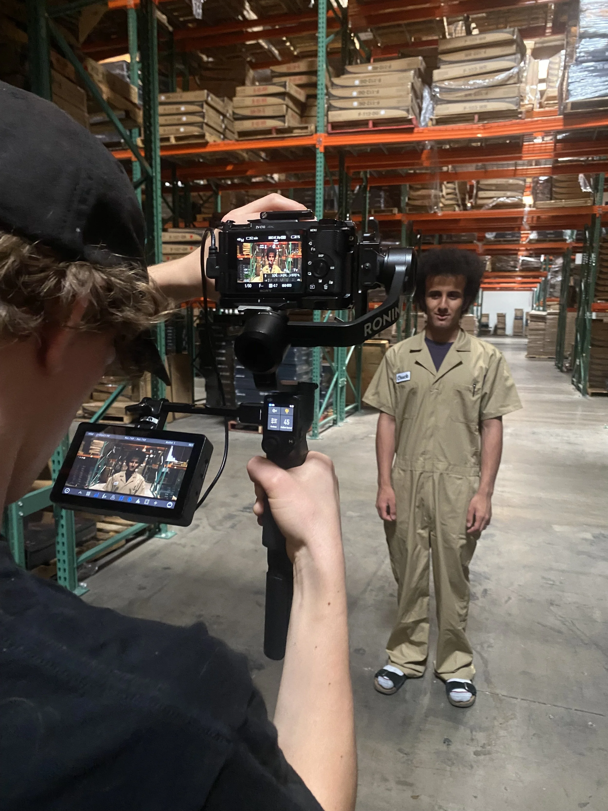 Film crew recording a worker in a warehouse aisle lined with stacked pallets and industrial shelving.