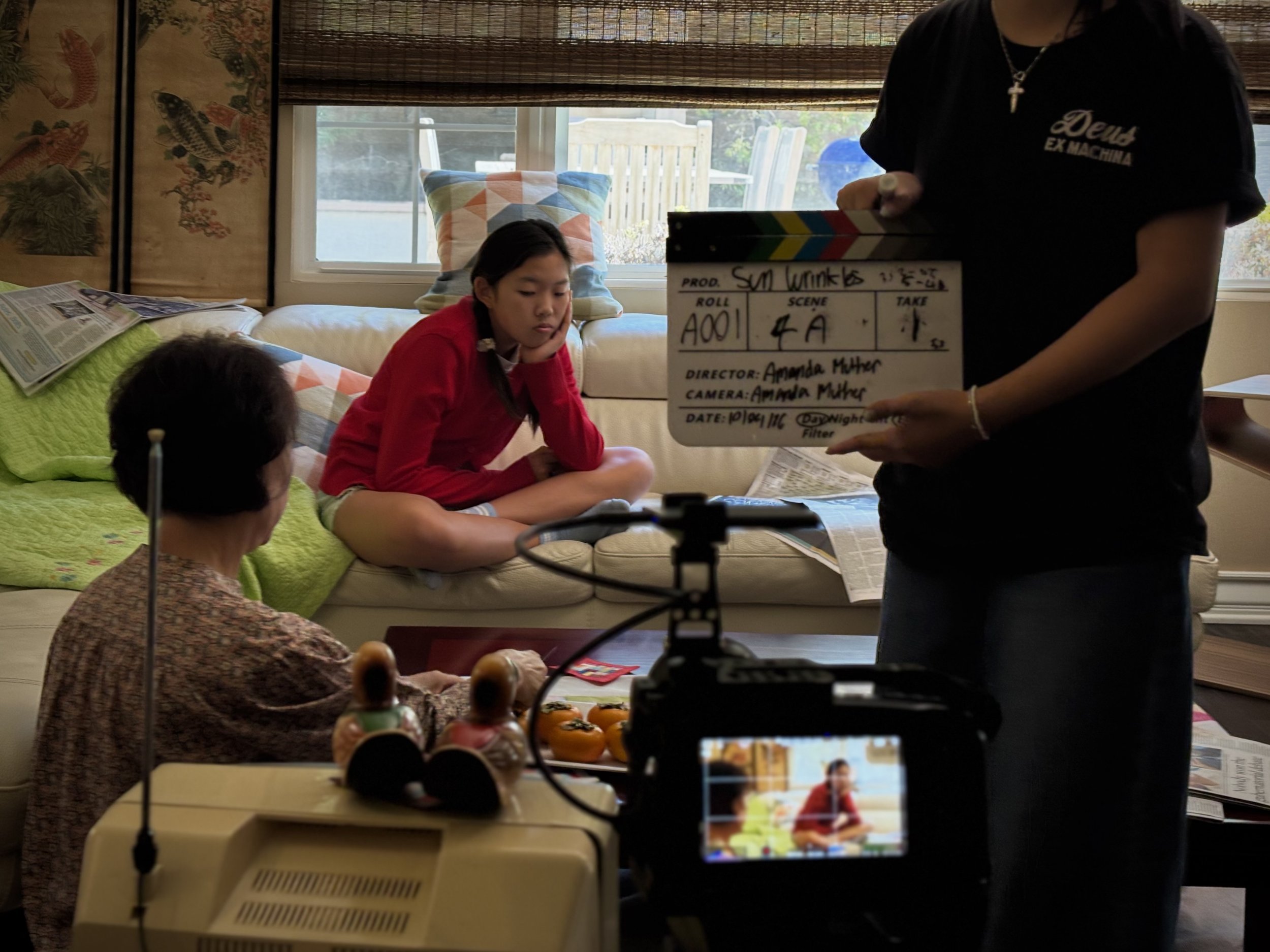 Film crew recording a young woman in a living room with a camera, monitor, and clapperboard.